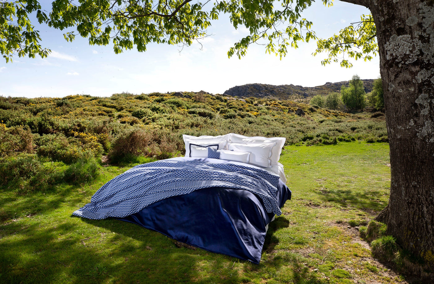 A bed with white pillows and blue patterned bedding is set up on a grassy meadow, featuring the Areo Pillowcase by Celso de Lemos available at Linen Society.