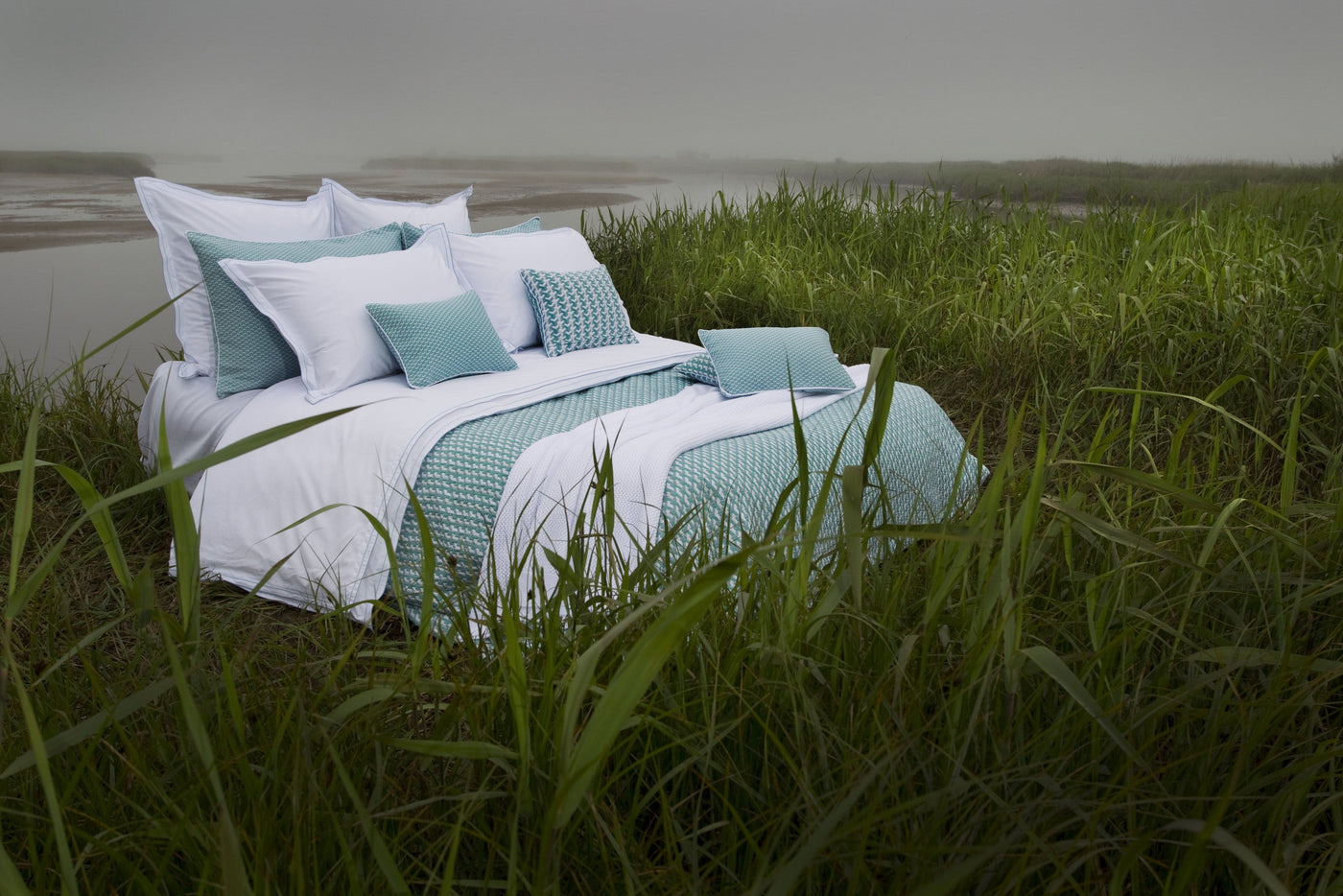 A bed with white linens and teal patterned pillows is set up outdoors in tall grass near a body of water, featuring Areo Pillowcase by Celso de Lemos available at Linen Society.