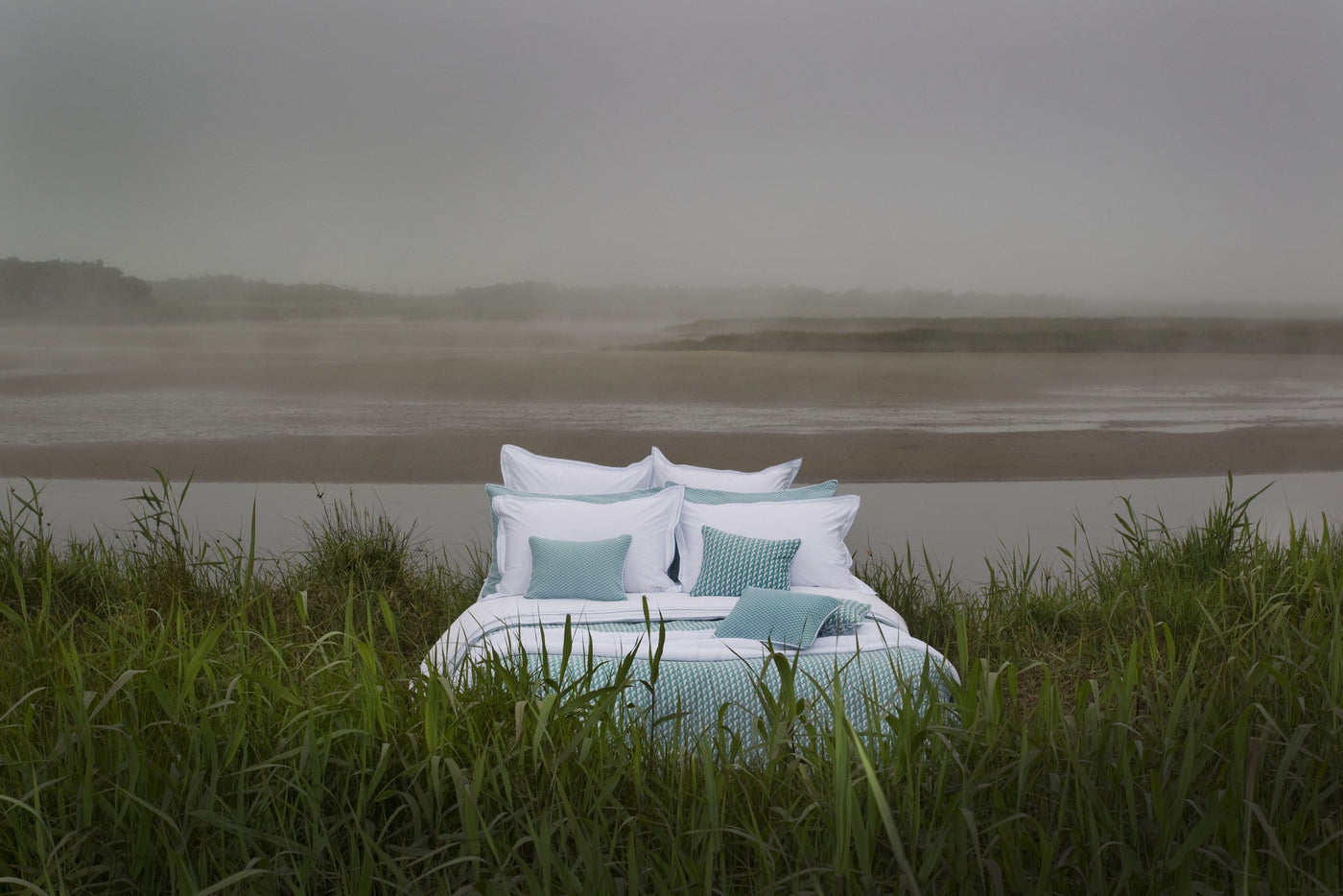 A neatly made bed with white bedding and teal accent pillows rests outdoors, surrounded by tall grass and a misty waterfront landscape, featuring the Areo Pillowcase by Celso de Lemos available at Linen Society.
