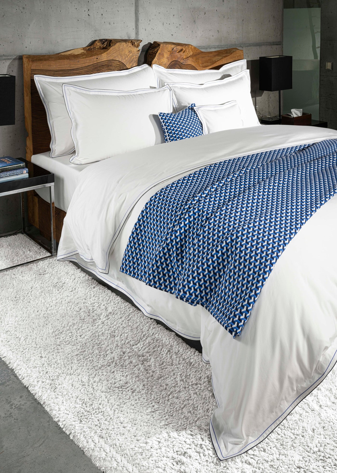Crisp white bedding with subtle blue piping and a geometric blue throw folded at the foot of the bed, featuring Areo Pillowcase by Celso de Lemos available at Linen Society.