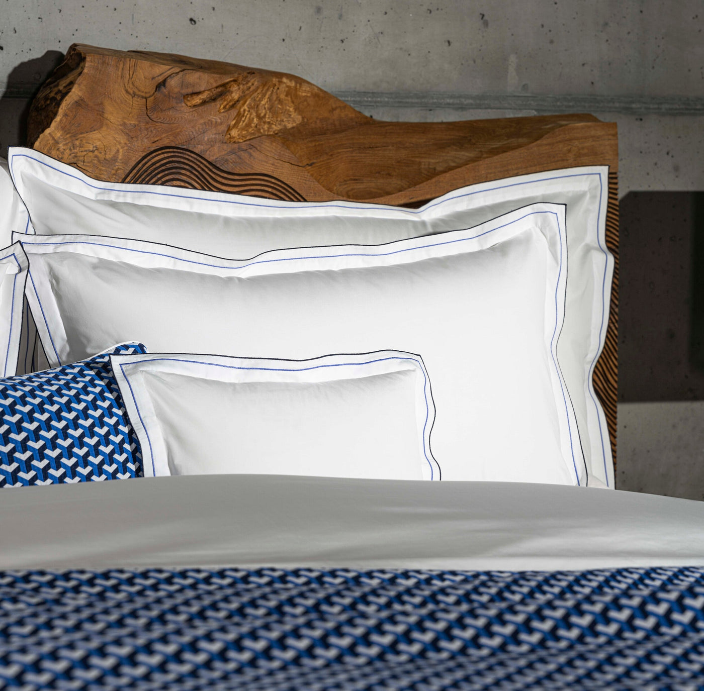 White Pillowcases with blue and black trim stacked neatly on a bed with a textured wooden headboard and a blue patterned throw blanket, featuring Areo Pillowcases by Celso de Lemos available at Linen Society.