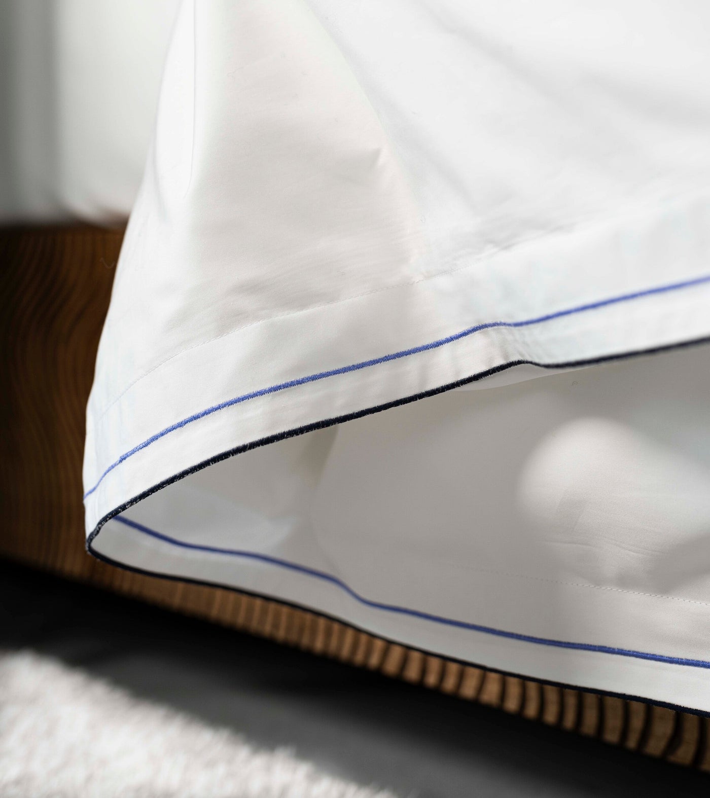 Close-up of white pillowcase with subtle blue and black piping, draped over a wooden headboard, featuring Areo Pillowcase by Celso de Lemos available at Linen Society.