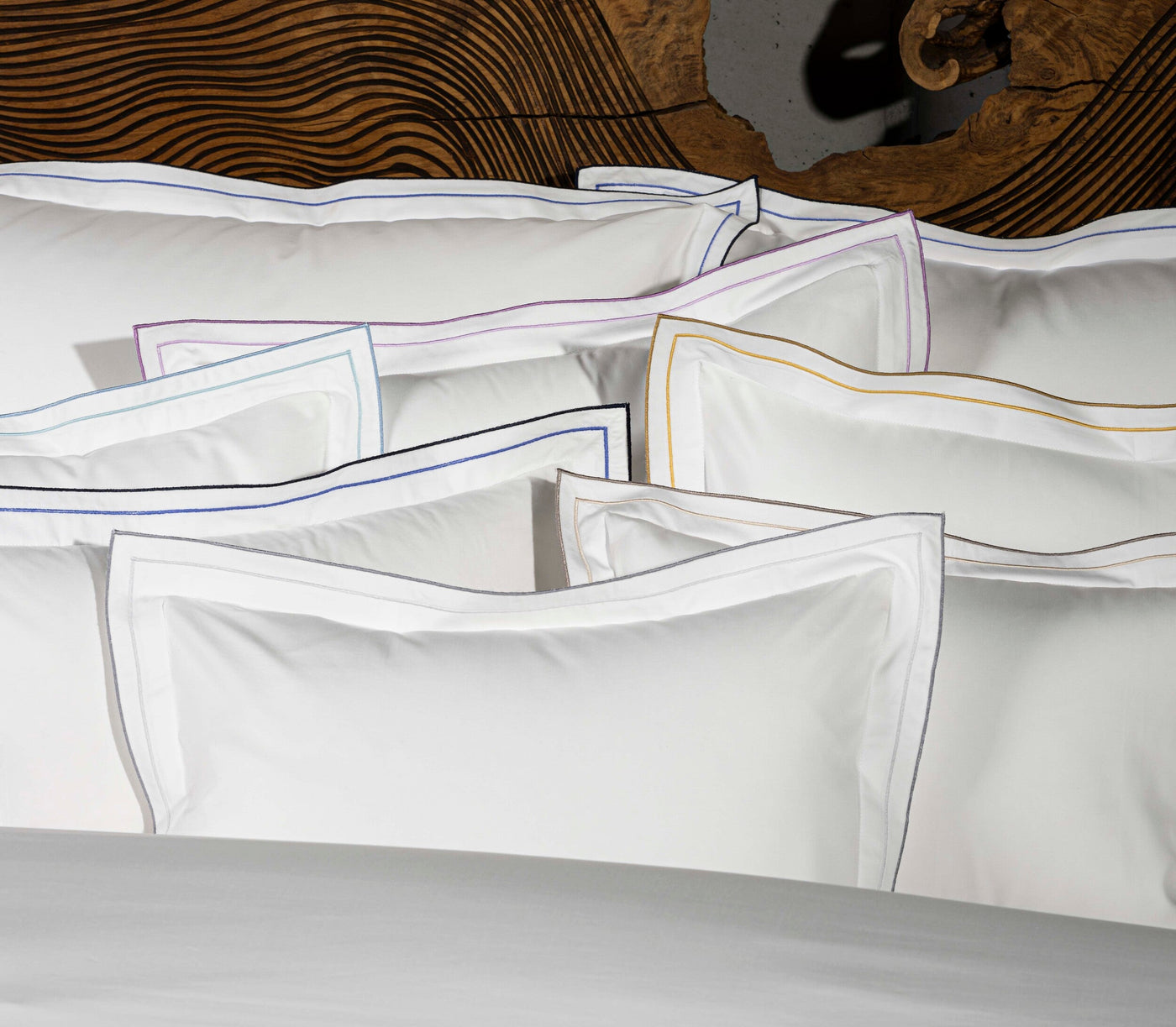 White pillowcases with contrasting colored piping, neatly stacked in front of a wooden headboard, featuring Areo Pillowcase by Celso de Lemos available at Linen Society.