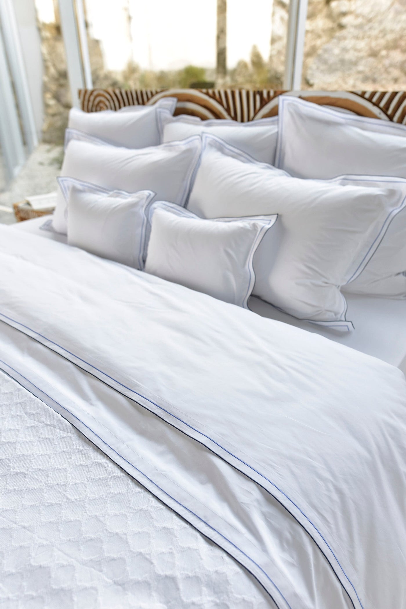 Crisp white bedding with subtle blue piping and a textured throw, featuring Areo Pillowcase by Celso de Lemos available at Linen Society.