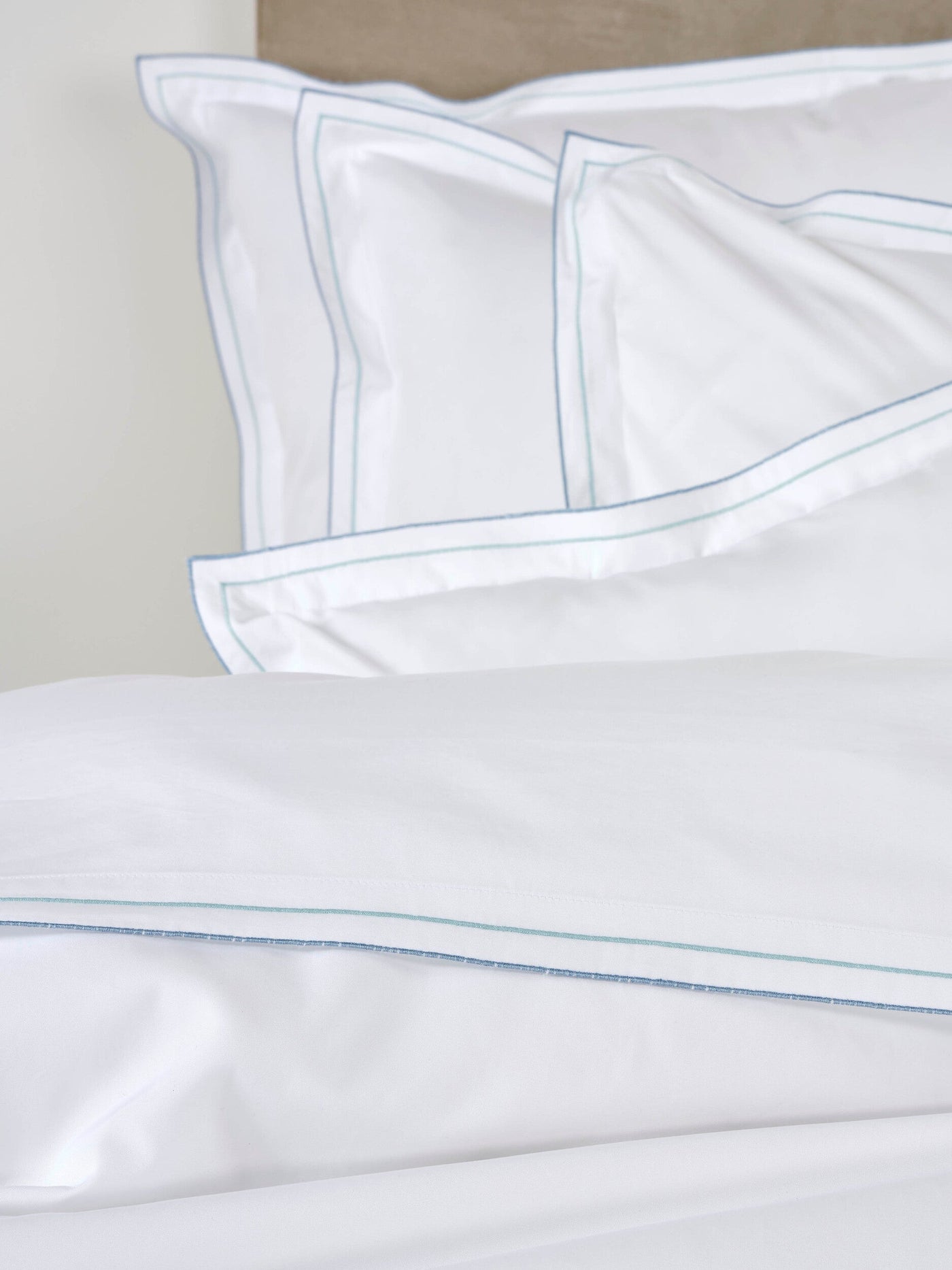 White pillowcases with fine blue and green stitching, neatly folded on a bed, featuring Areo Pillowcase by Celso de Lemos available at Linen Society.