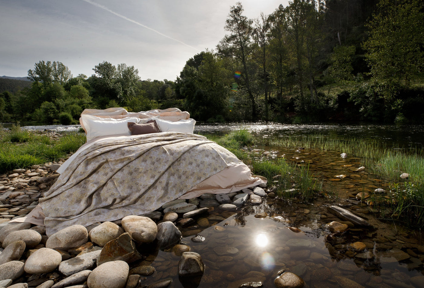 White pillow covers and a patterned duvet on a bed, set on a rocky riverbank by a river with trees in the background, featuring Australia Pillow Cover by Celso de Lemos available at Linen Society.