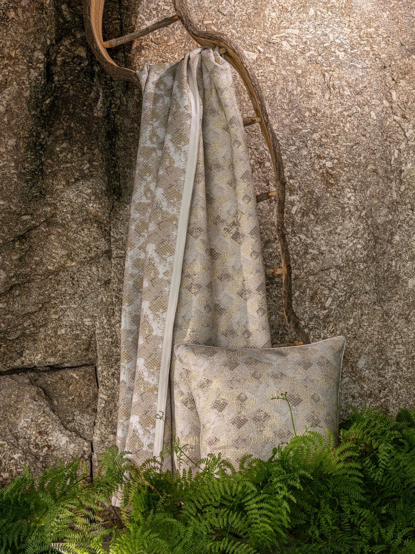 A neutral-toned pillow and draped bed cover with a subtle snake-print texture and a gold shimmer, resting against a rustic wooden ladder and a textured rock face, surrounded by lush green ferns, featuring Australia Bed Cover by Celso de Lemos available at Linen Society.