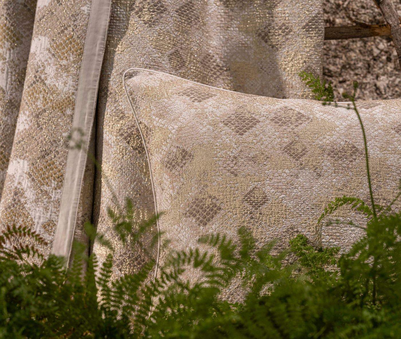 Close-up of a shimmering gold and beige textured Pillow Cover with a subtle raised pattern, partially visible behind green ferns. featuring Australia Pillow Cover by Celso de Lemos available at Linen Society.