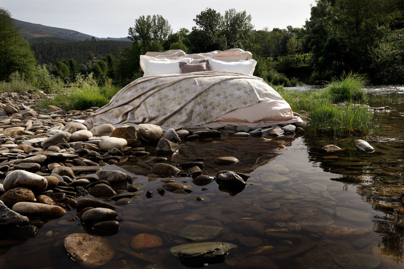 A bed with smooth white and light brown pillow covers and a patterned gold duvet set on river rocks with water and green trees. featuring Australia Pillow Cover by Celso de Lemos available at Linen Society.