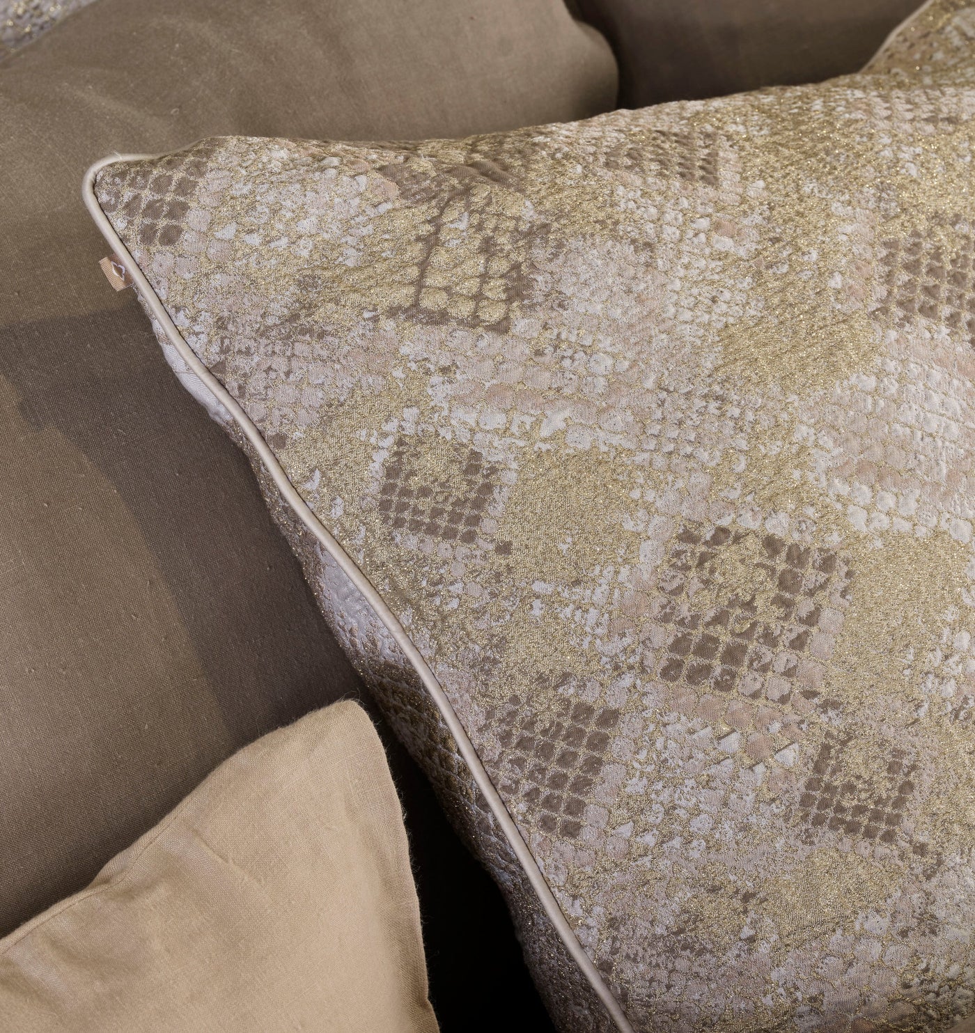 Close-up of a beige and gold metallic jacquard patterned pillow with piped edges, resting on solid neutral cushions. featuring Australia Pillow Cover by Celso de Lemos available at Linen Society.