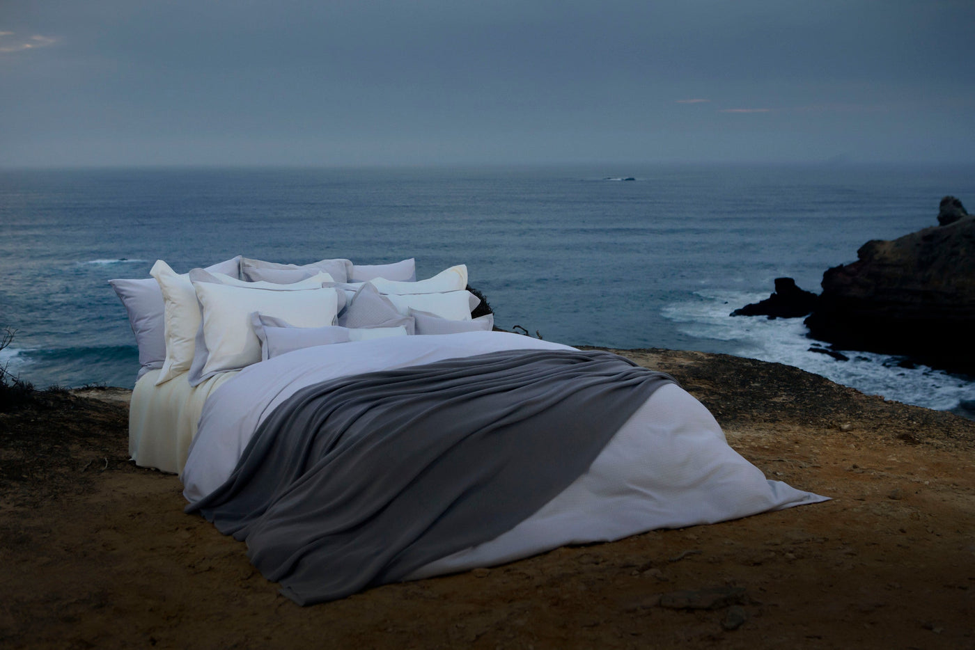 A bed dressed in white and grey linens is set against the backdrop of a serene ocean view. featuring Avalon Pillowcase by Celso de Lemos available at Linen Society.