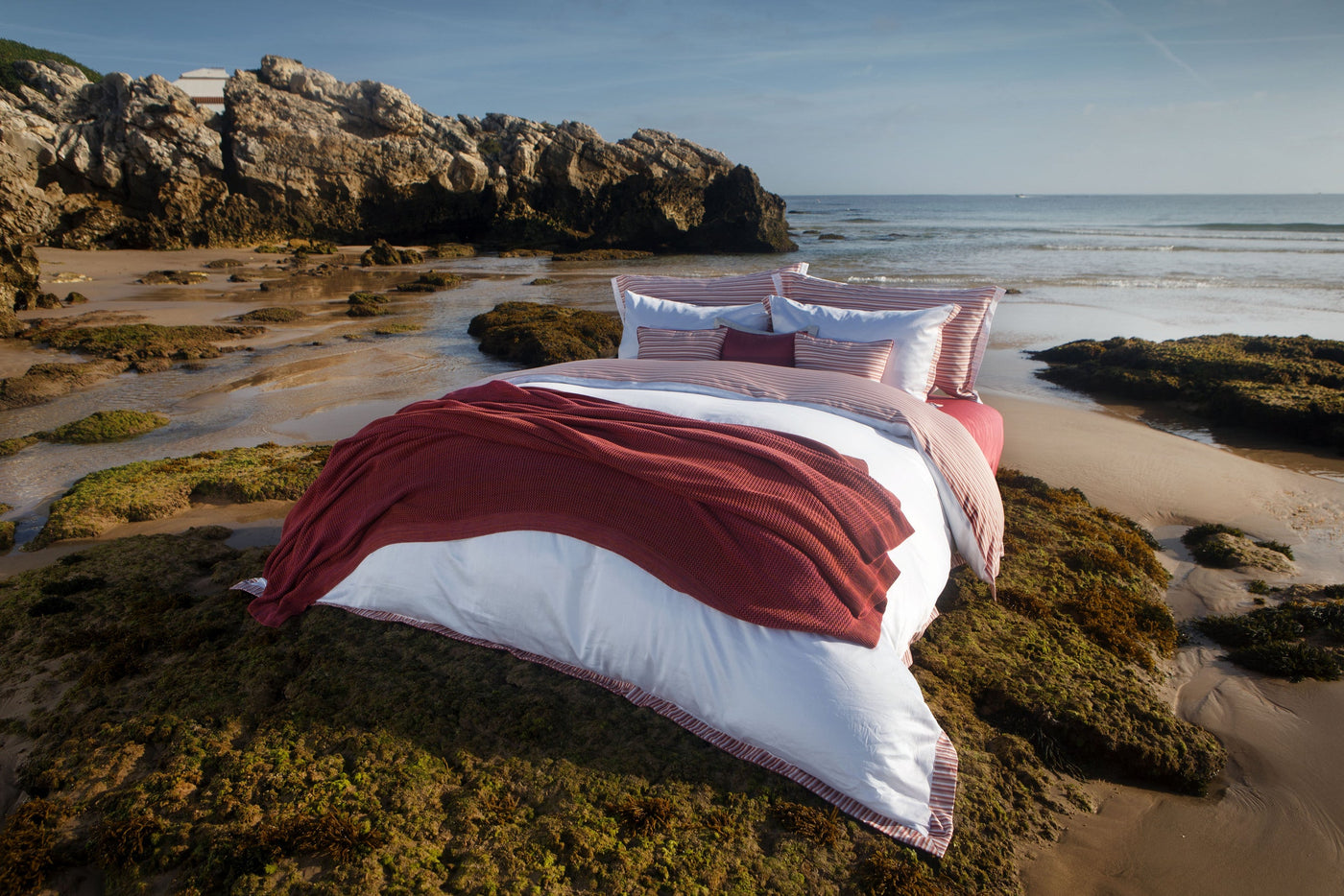 A bed with white and striped linens and a burgundy throw is set on a rocky beach. featuring Baya Pillowcase by Celso de Lemos available at Linen Society.