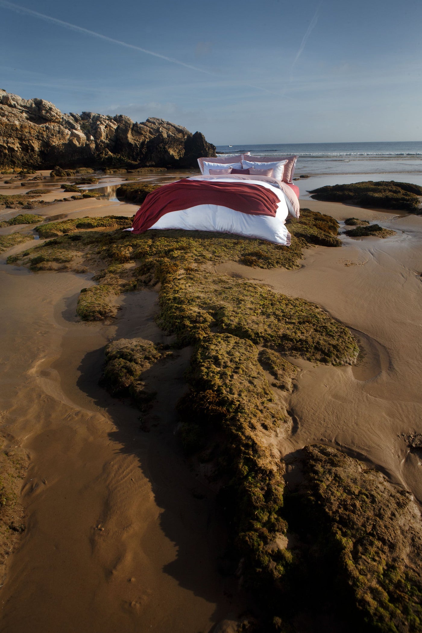 A bed with white and red linens is displayed on a rocky beach with the ocean in the background, featuring Baya Pillowcase by Celso de Lemos available at Linen Society.