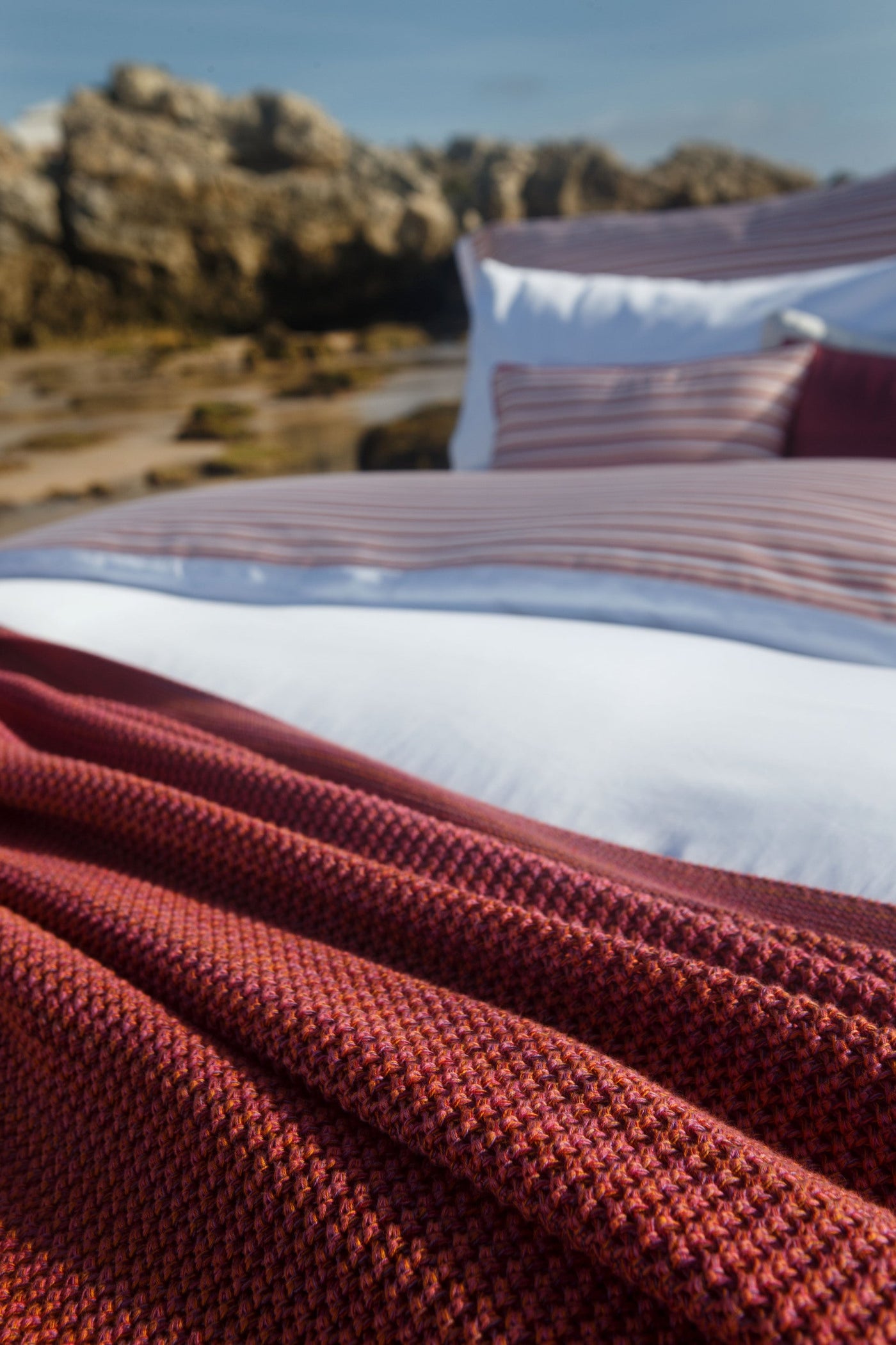 A textured crimson knit throw is draped over a bed with striped and white linens, set against a backdrop of rocky coastal terrain. featuring Baya Pillowcase by Celso de Lemos available at Linen Society.