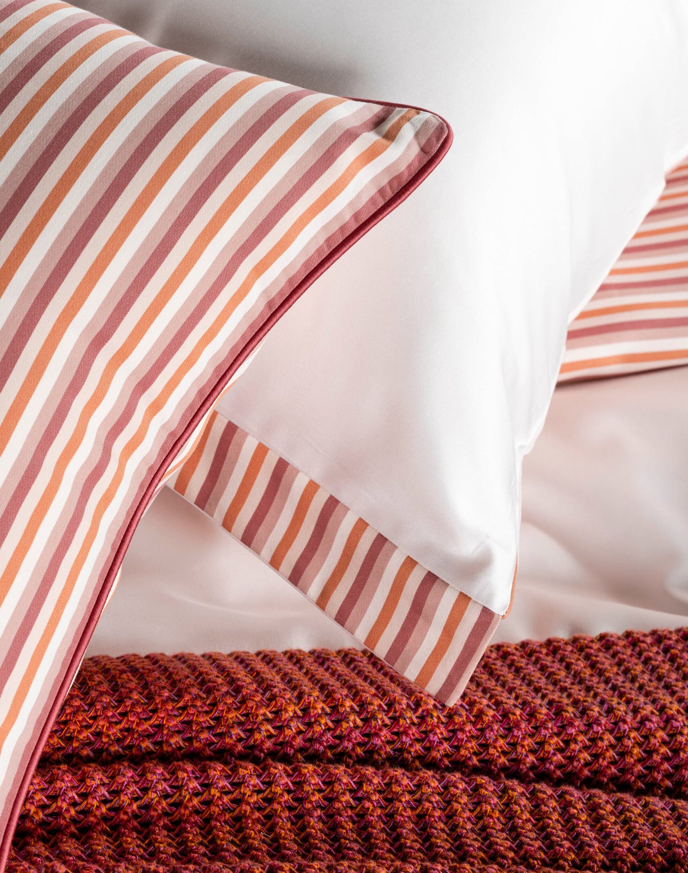 Close-up of a bed featuring striped pillowcases in shades of rose, peach, and cream, with a textured rust-colored throw, featuring Baya Pillowcase by Celso de Lemos available at Linen Society.