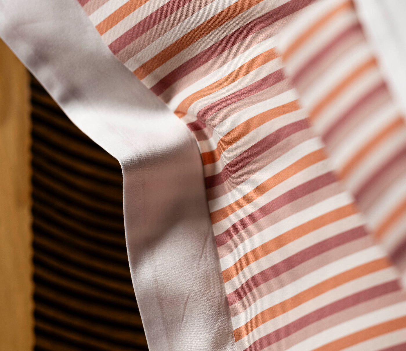 Close-up of striped pillowcase fabric in muted pinks, oranges, and cream, with a satin trim. featuring Baya Pillowcase by Celso de Lemos available at Linen Society.