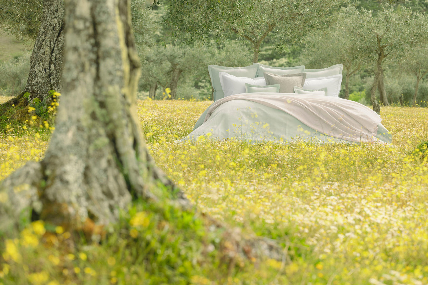 A bed with light green bedding and a blush pink throw is set in a sunny field of wildflowers, featuring Bijoux Pillowcases by Celso de Lemos available at Linen Society.
