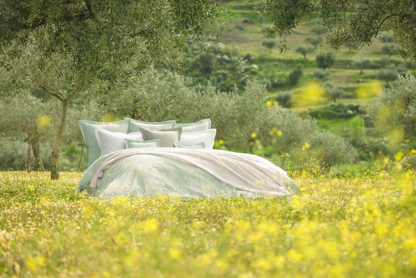 A bed with pale green bedding and white pillows is set outdoors amongst olive trees and a field of yellow wildflowers, featuring Bijoux Pillowcase by Celso de Lemos available at Linen Society.
