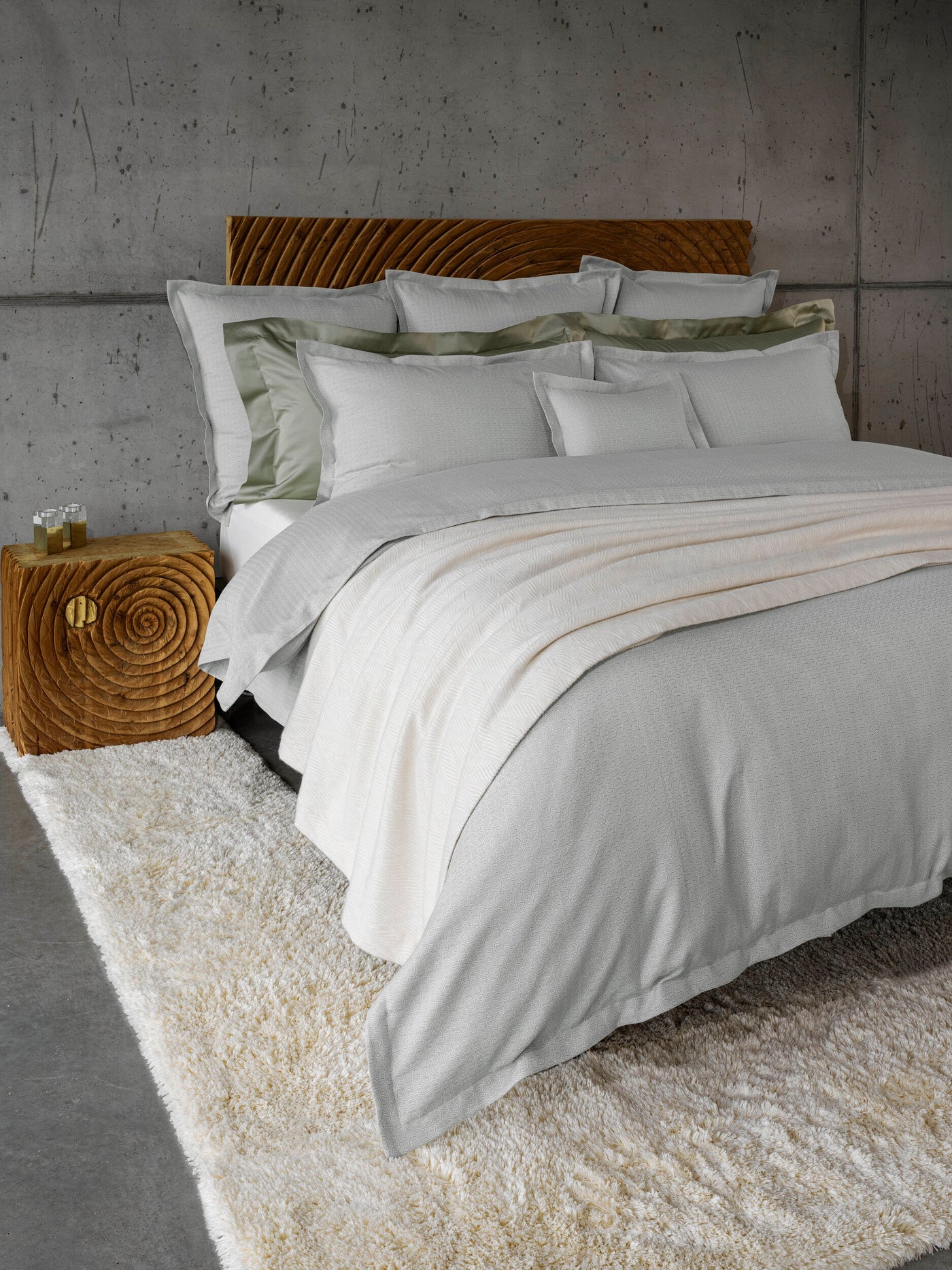 A neatly made bed with white and sage green bedding is layered with a white textured throw, set against a concrete wall and a textured wooden nightstand. featuring Bijoux Pillowcase by Celso de Lemos available at Linen Society.
