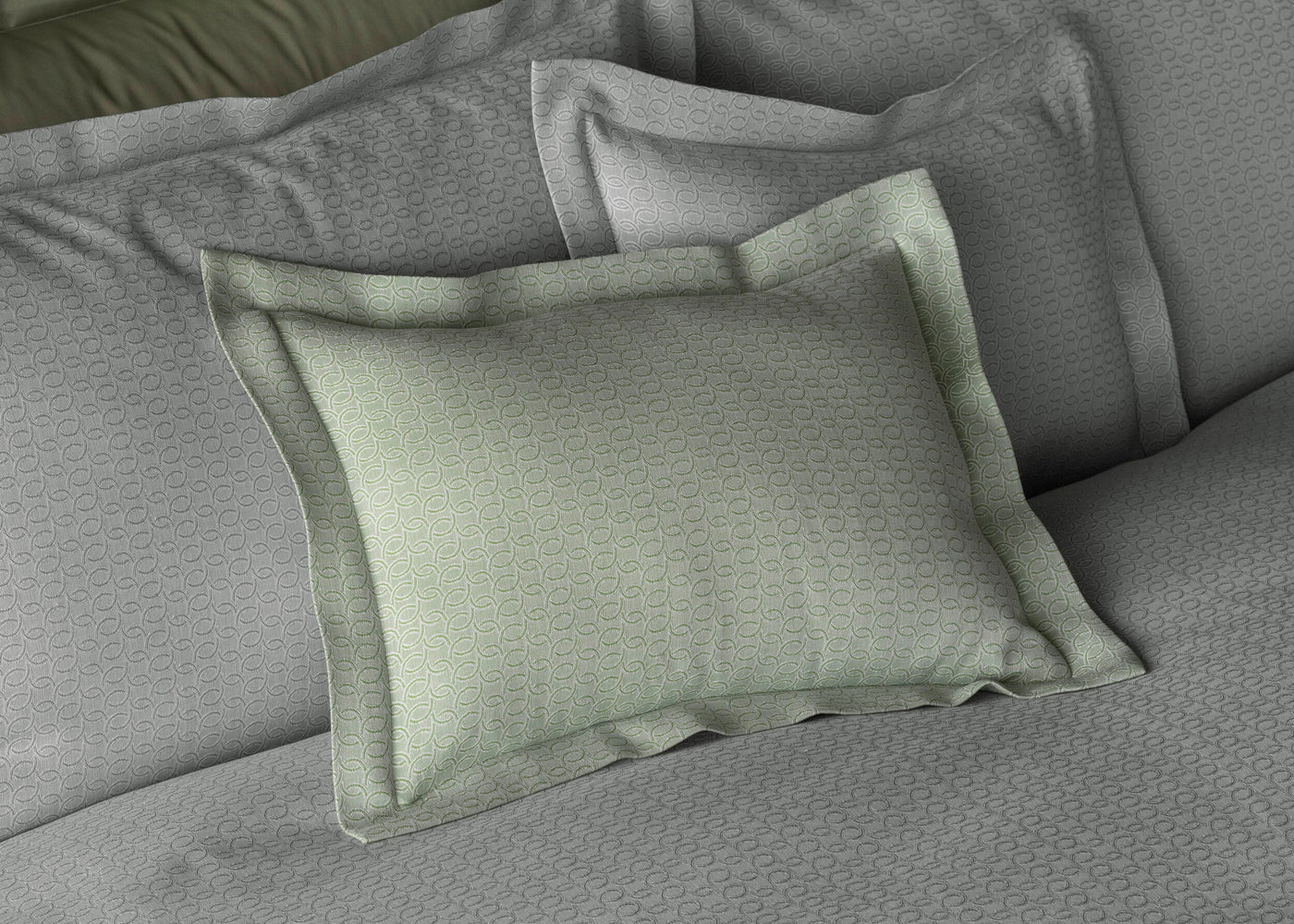 A light green pillowcase with a subtle swirling pattern is layered on a bed with grey bedding. The fabric appears soft and textured. featuring Bijoux Pillowcase by Celso de Lemos available at Linen Society.