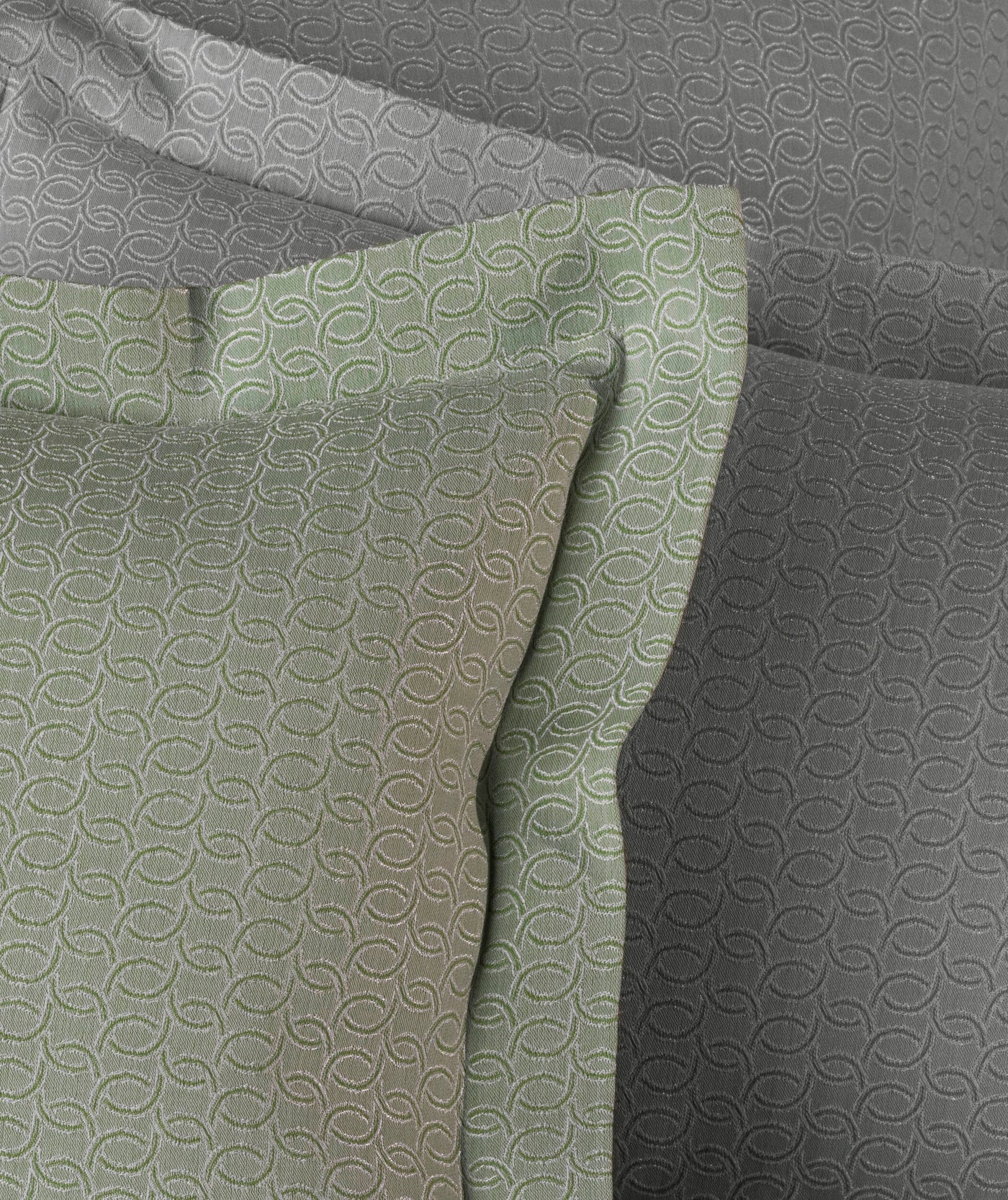 A sage green pillowcase with a textured interlocking ring pattern lies atop grey pillowcases featuring a similar subtle pattern. featuring Bijoux Pillowcase by Celso de Lemos available at Linen Society.