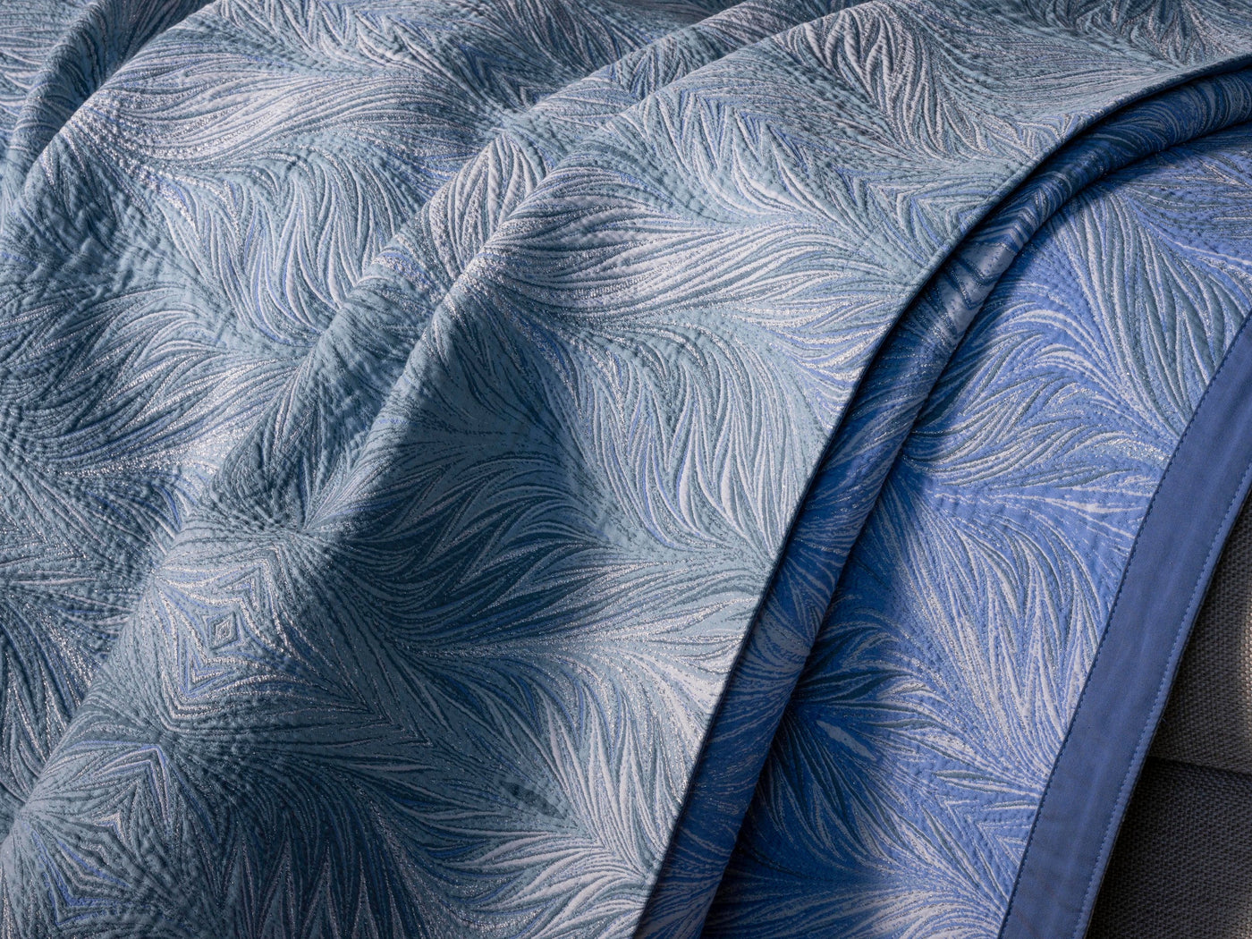 Close-up of light blue textured fabric with a shimmering feather pattern and a darker blue border, featuring Boheme Bed Cover by Celso de Lemos available at Linen Society.