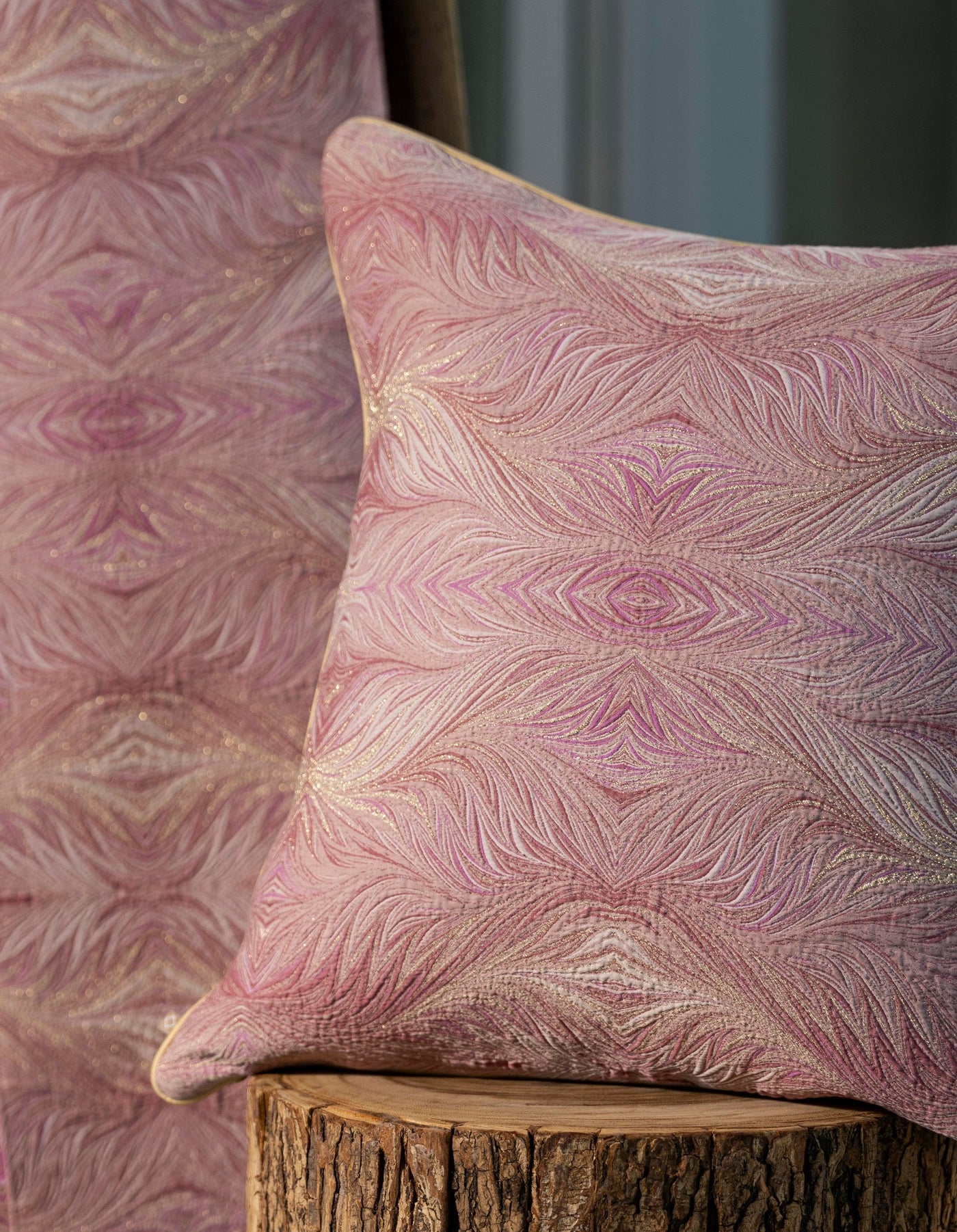 A pink pillow with a feathered pattern and subtle shimmer rests on a wooden stump, with a matching fabric behind it, featuring Boheme Pillow Cover by Celso de Lemos available at Linen Society.