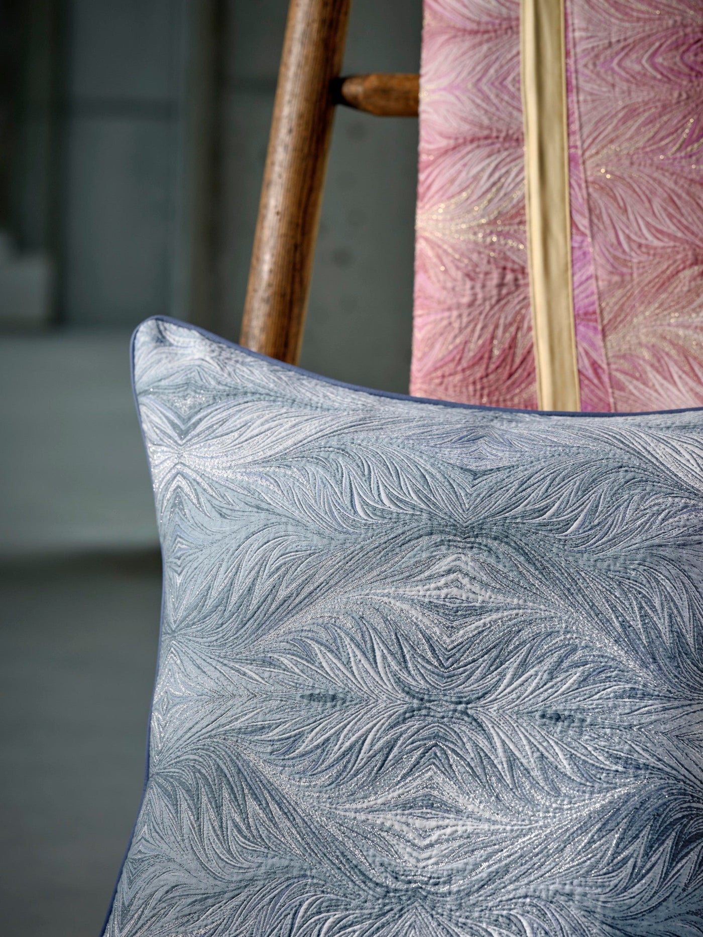 A textured blue pillow cover with a swirling pattern and metallic threads is displayed on a wooden chair next to a pink textile. featuring Boheme Pillow Cover by Celso de Lemos available at Linen Society.