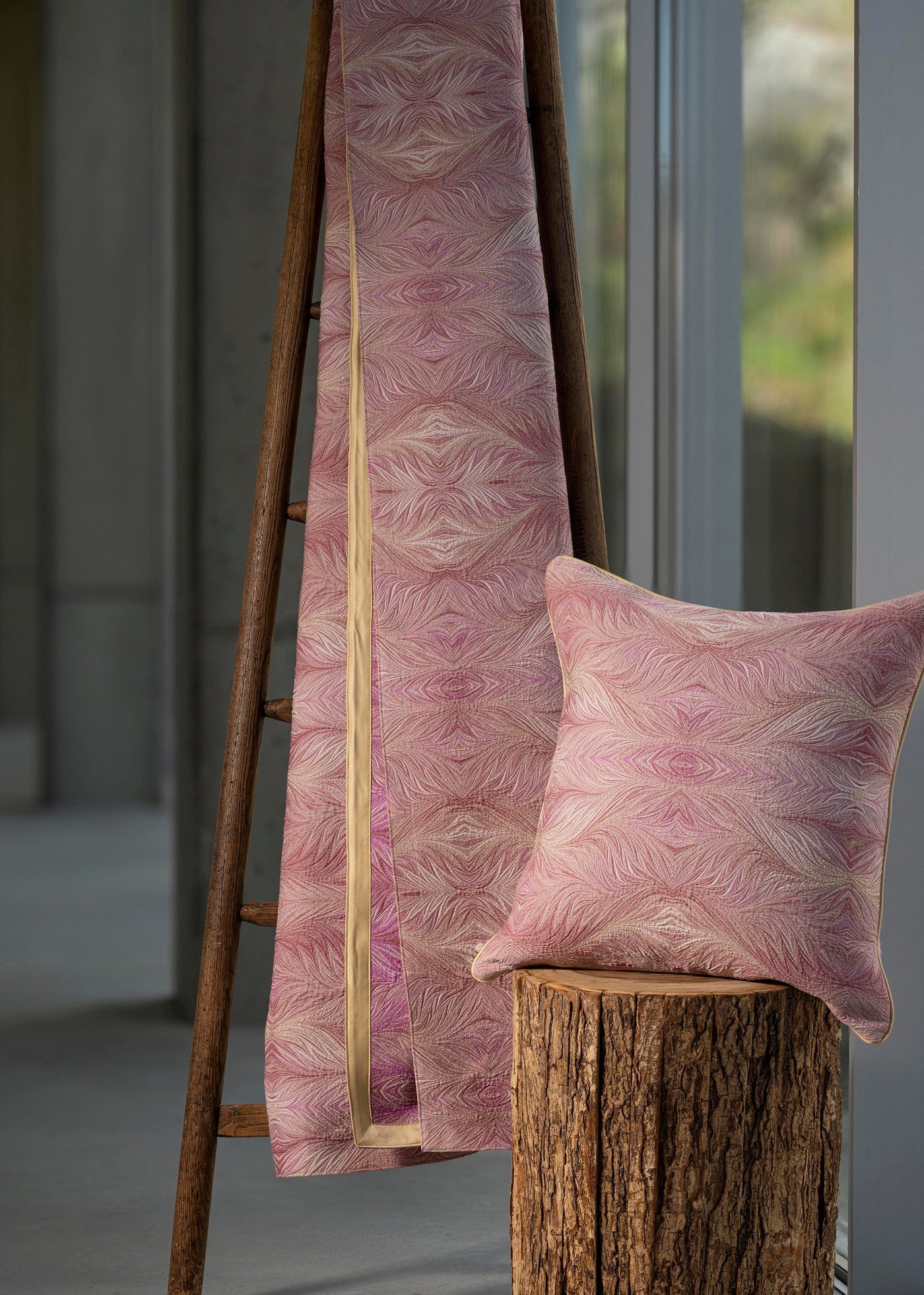 A pink pillow and blanket with a subtle shimmer feature an abstract pattern, displayed on a wooden ladder and tree stump. featuring Boheme Pillow Cover by Celso de Lemos available at Linen Society.