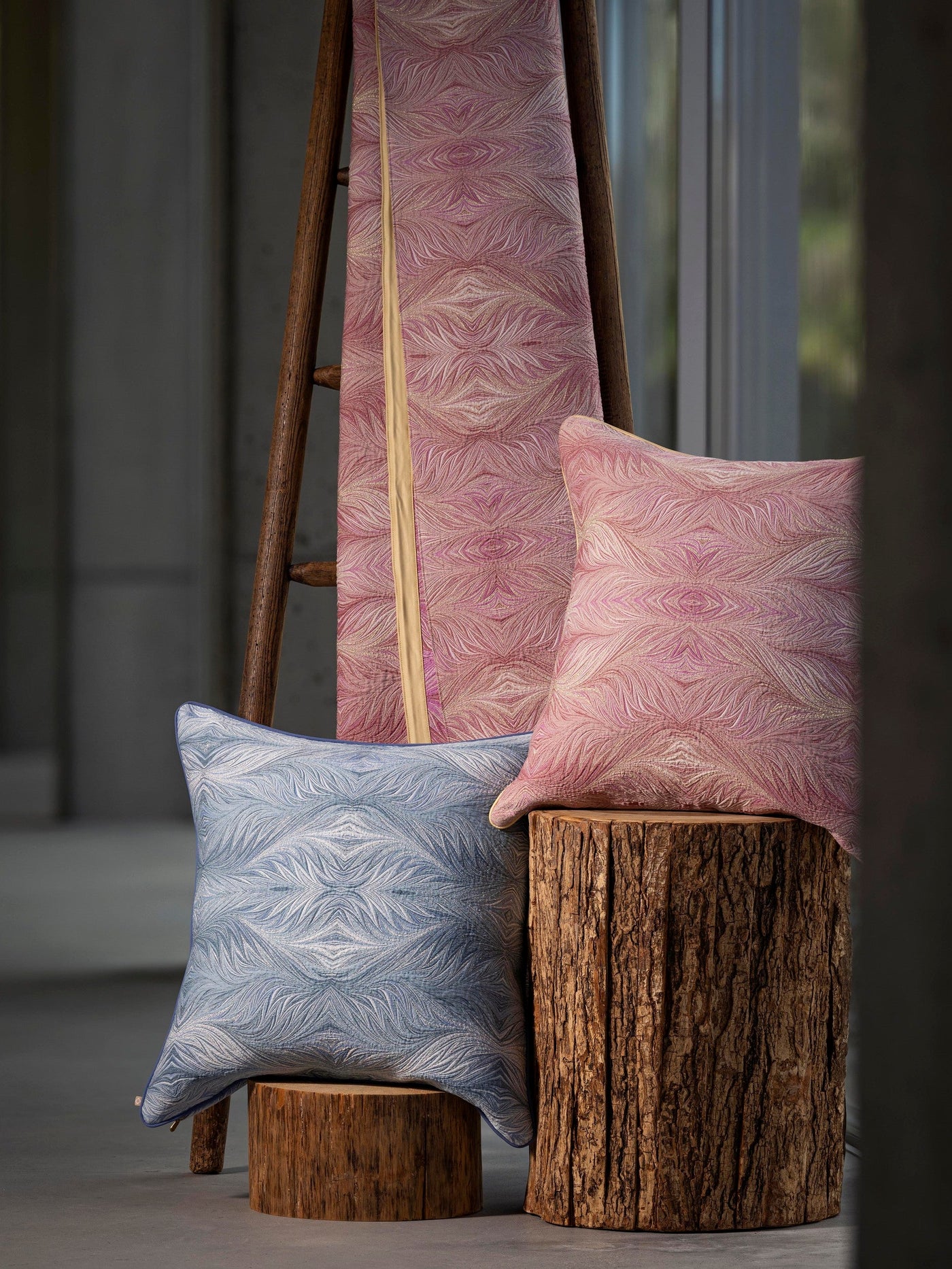 Two textured pillow covers, one in blue and one in pink, are displayed near a wooden ladder and on log stumps, featuring Boheme Pillow Cover by Celso de Lemos available at Linen Society.