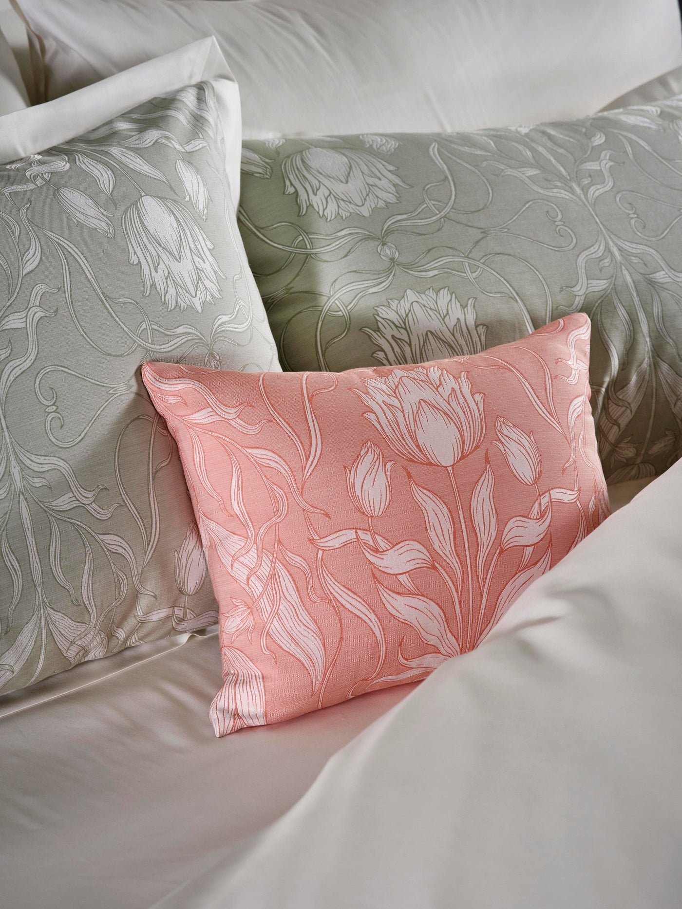 A close-up of a bed with several pillows showcasing a floral pattern. One pillow is coral-pink with white floral motifs, while others are sage-green. A light-colored duvet is visible. featuring Botanica Pillowcase by Celso de Lemos available at Linen Society.