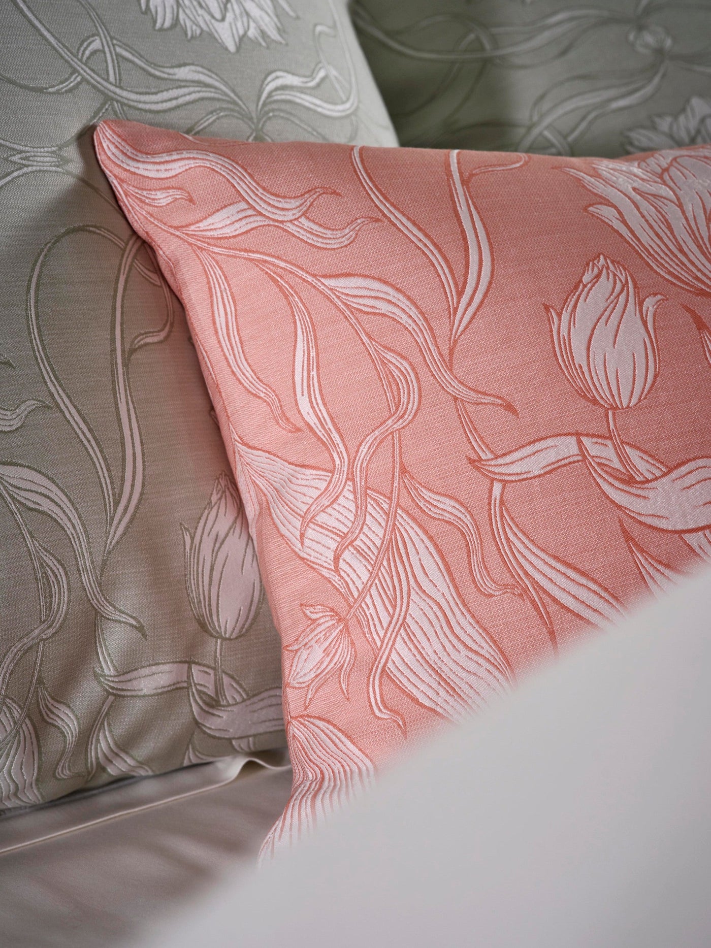 Close-up of two textured pillowcases, one in coral pink and one in muted green, both with intricate white floral patterns. featuring Botanica Pillowcase by Celso de Lemos available at Linen Society.