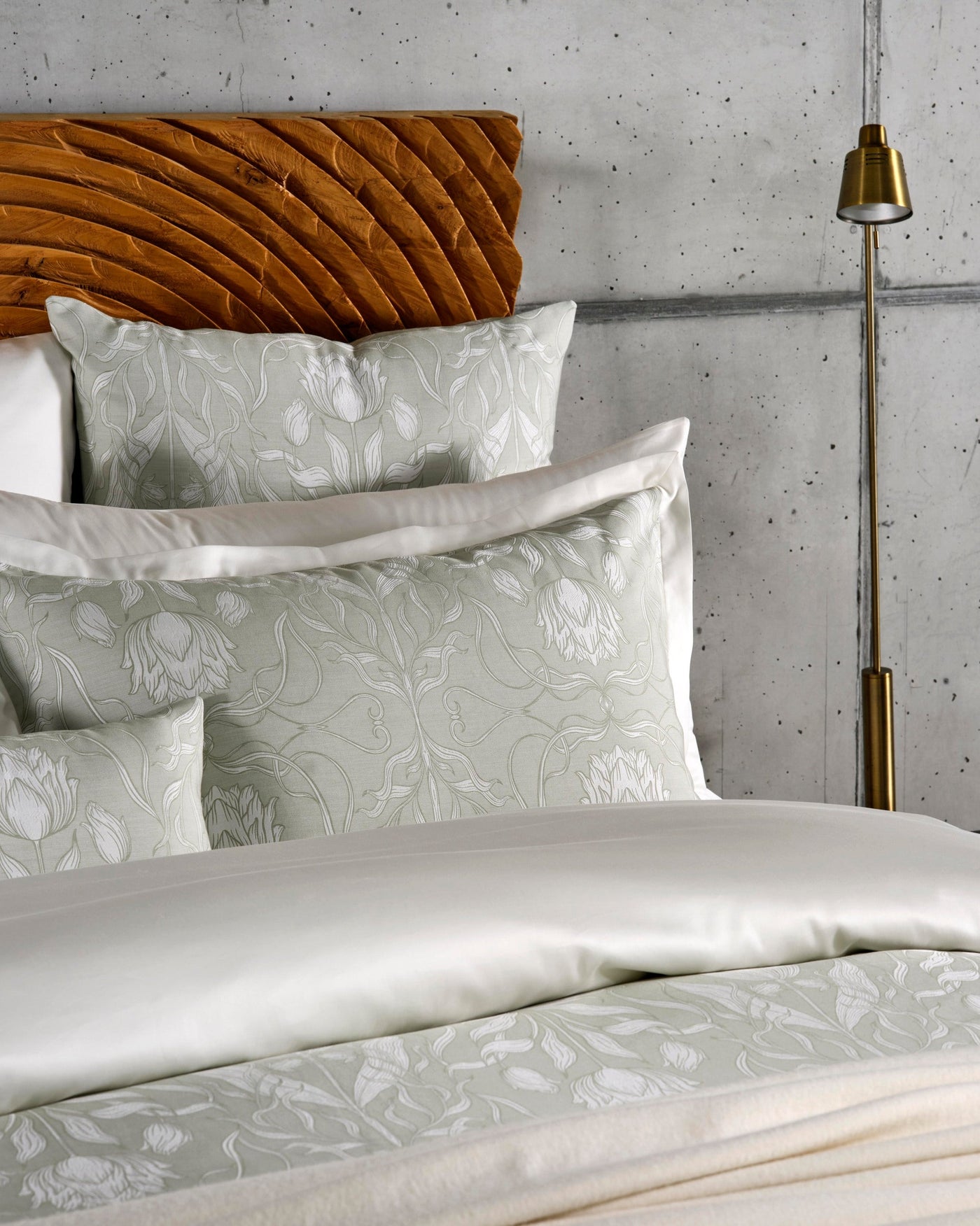 A bed with textured pillows featuring a white floral pattern on a light green background, set against a concrete wall and wooden headboard, featuring Botanica Pillowcase by Celso de Lemos available at Linen Society.