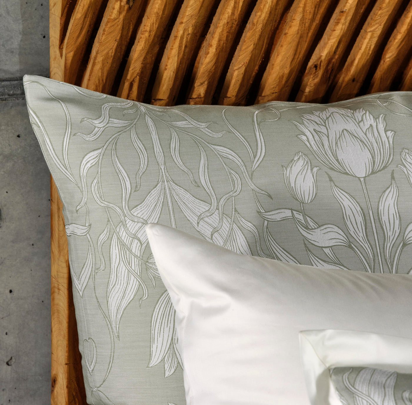 Close-up of a light green pillowcase with white floral embroidery, placed on a bed with a textured wooden headboard and a white pillow. featuring Botanica Pillowcase by Celso de Lemos available at Linen Society.