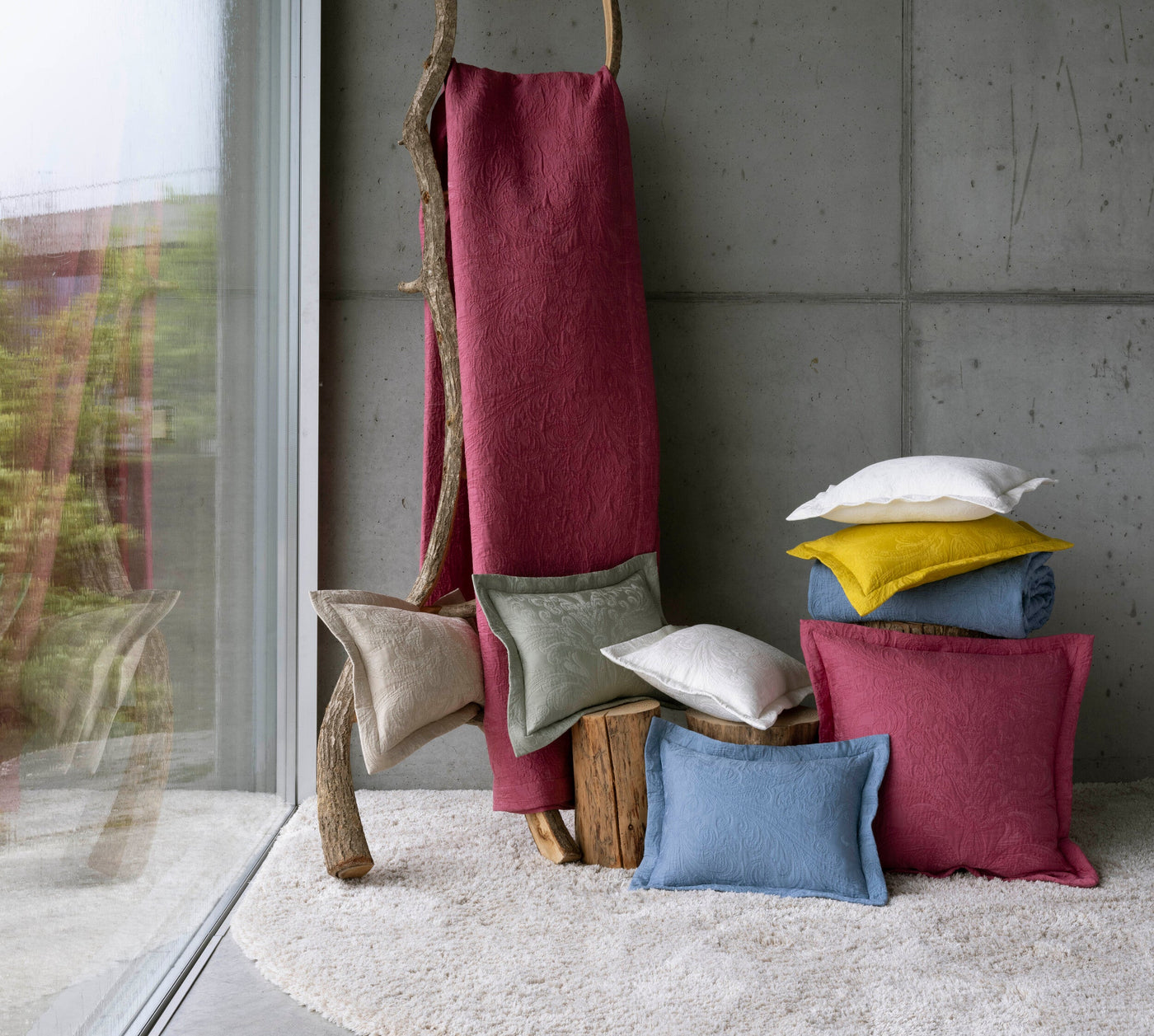 Pillows and a draped textile with embossed floral pattern in berry, blue, sage green, natural, yellow, and white are arranged on a textured rug and wooden logs. featuring Braga Pillow Cover by Celso de Lemos available at Linen Society.
