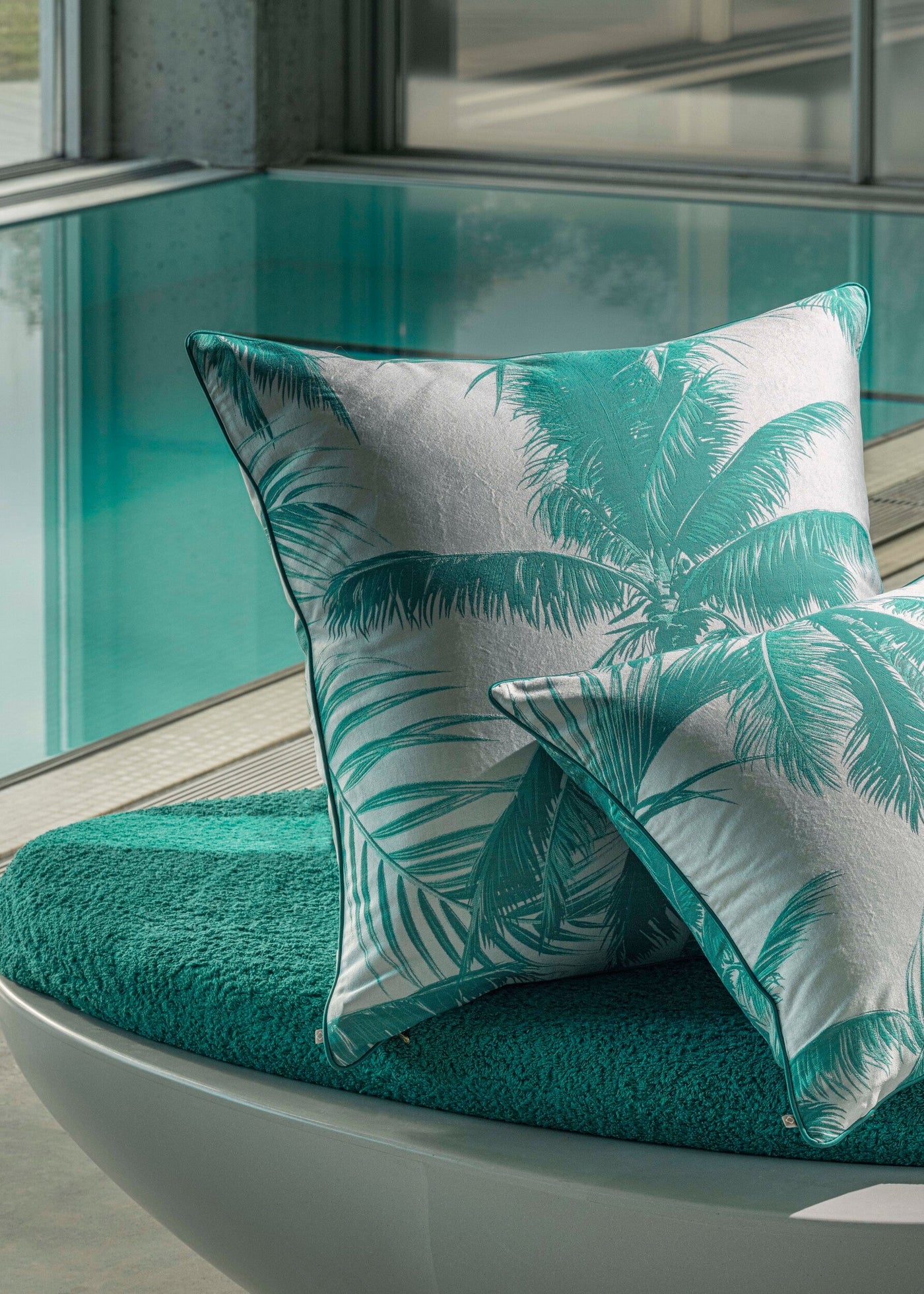 Two white pillows with a teal palm frond pattern rest on a textured teal cushion by a pool, featuring Cabo Verde Pillow Cover by Celso de Lemos available at Linen Society.