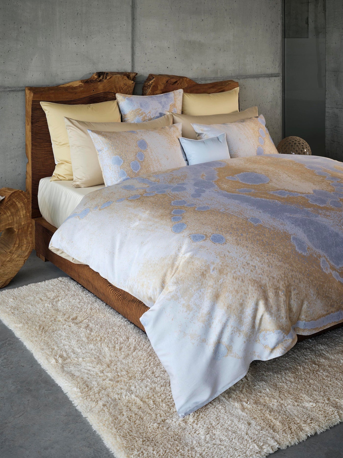 A bed with a textured wooden headboard and bedding with a marbled pattern in gold and blue tones, featuring Callisto Pillowcase by Celso de Lemos available at Linen Society.