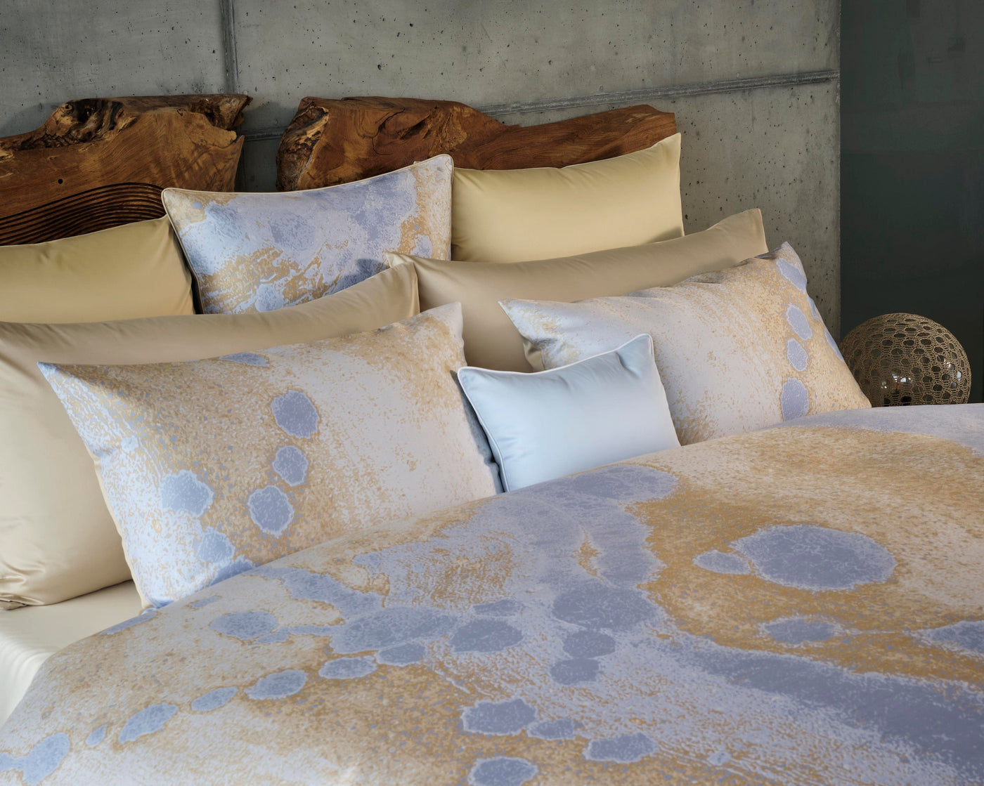 A bed with a textured concrete wall and a wooden headboard, adorned with soft, light-colored bedding featuring a subtle abstract pattern of blue and gold. Featuring Callisto Pillowcase by Celso de Lemos available at Linen Society.