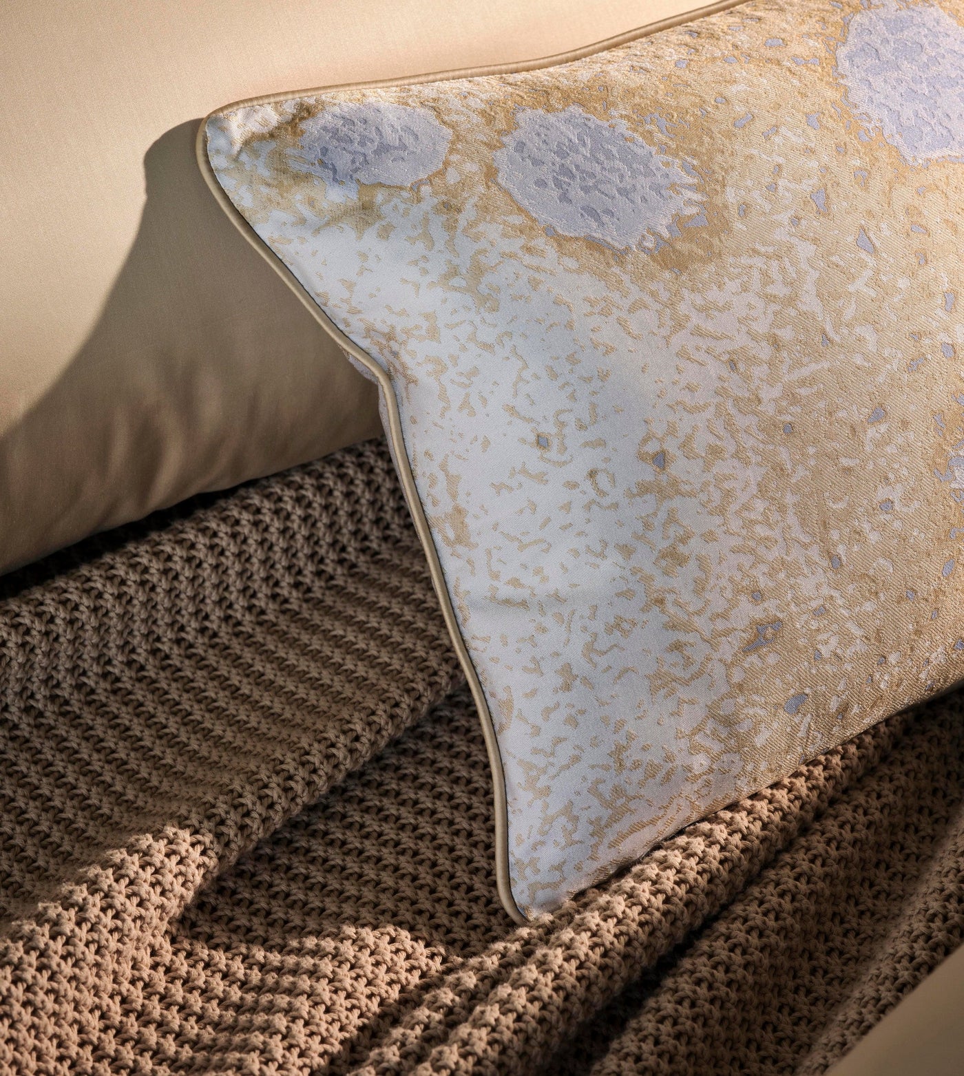 A textured knit throw is layered beneath a decorative pillow with a subtle abstract pattern and gold piping. featuring Callisto Pillowcase by Celso de Lemos available at Linen Society.