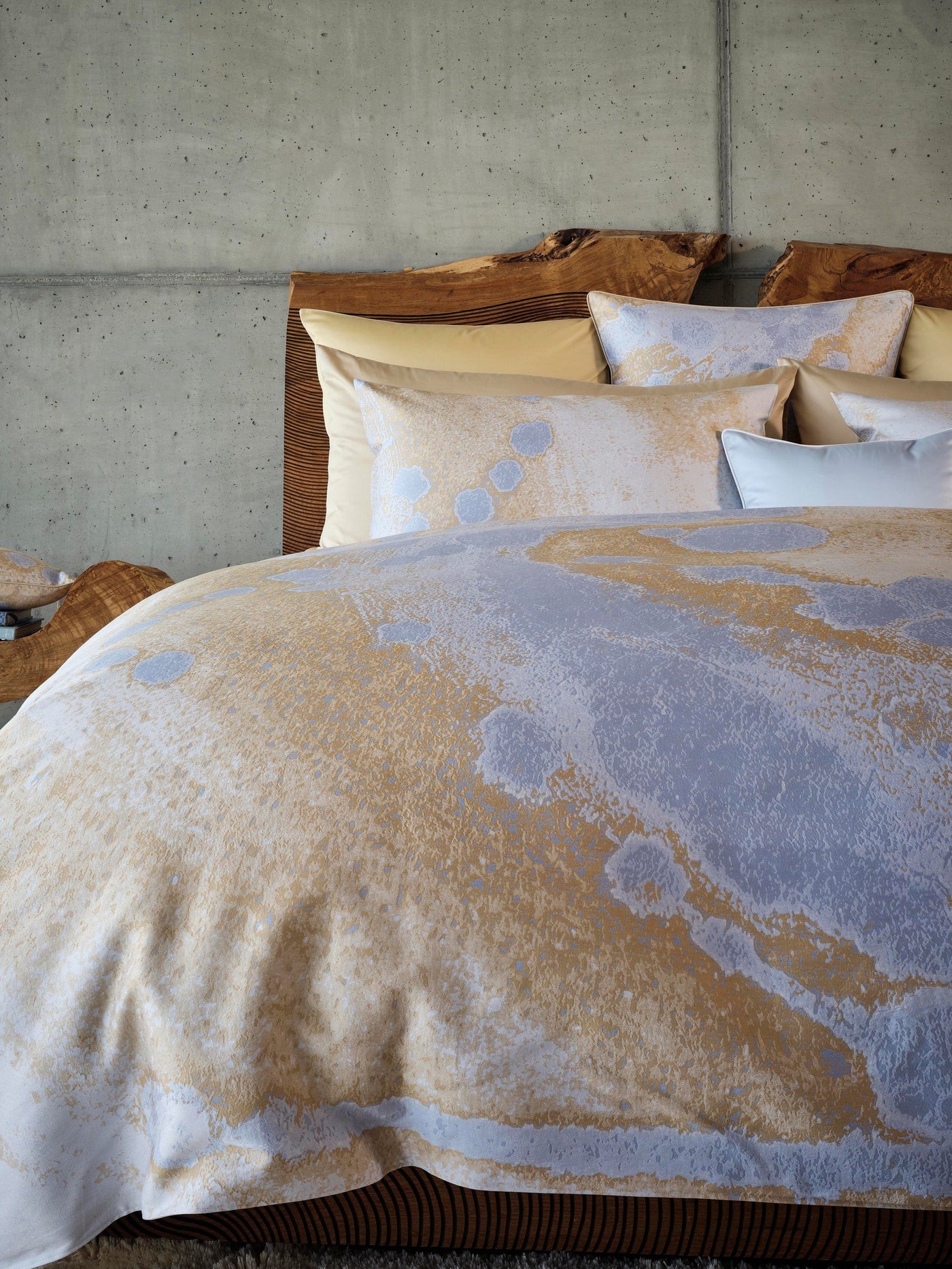 Bedding with a textured abstract pattern in gold and grey on a concrete background, featuring the Callisto Pillowcase by Celso de Lemos available at Linen Society.