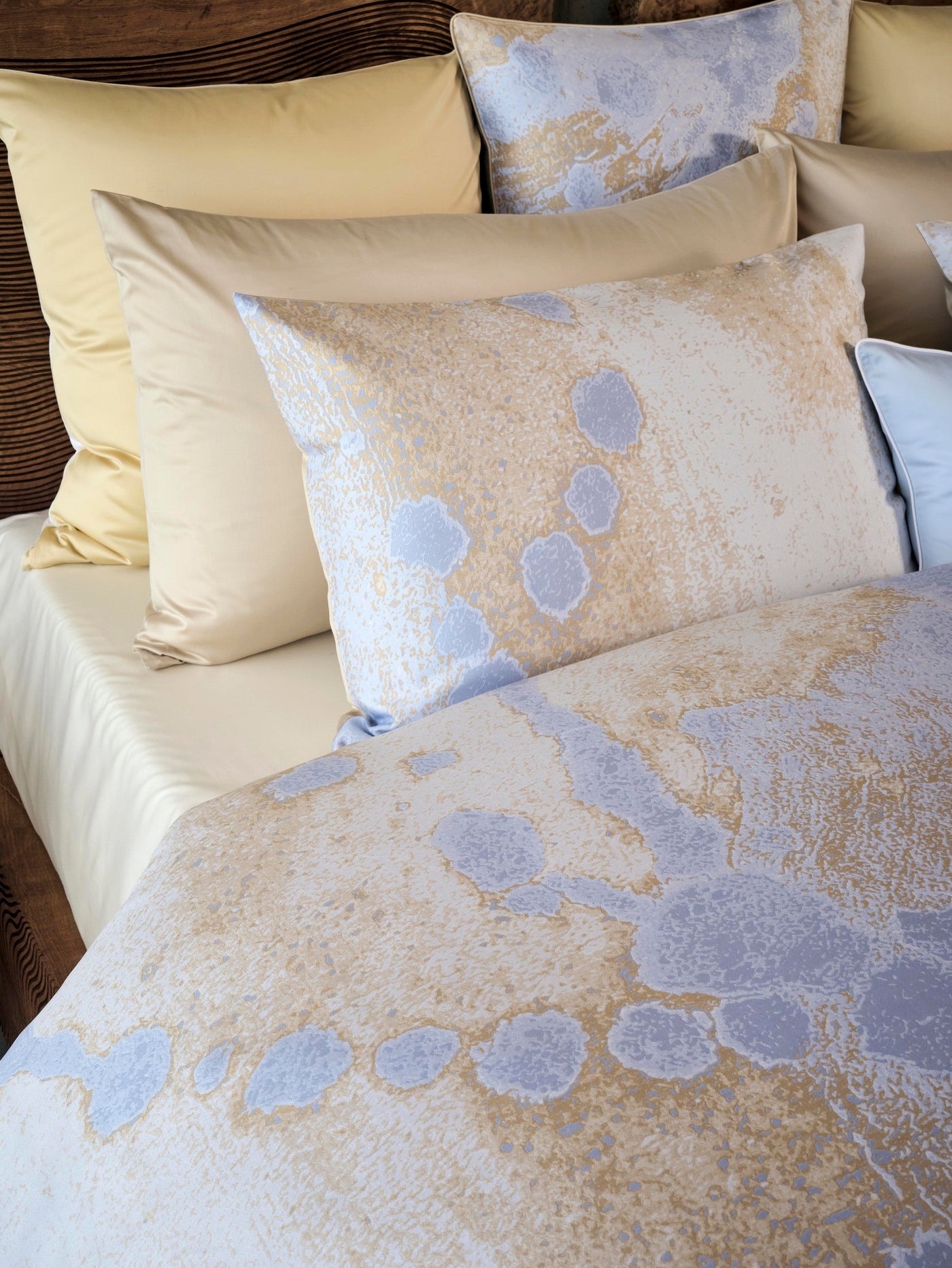 A close-up of a bed with layered pillows and a duvet featuring a blue and beige abstract splatter pattern. Featuring Callisto Pillowcase by Celso de Lemos available at Linen Society.