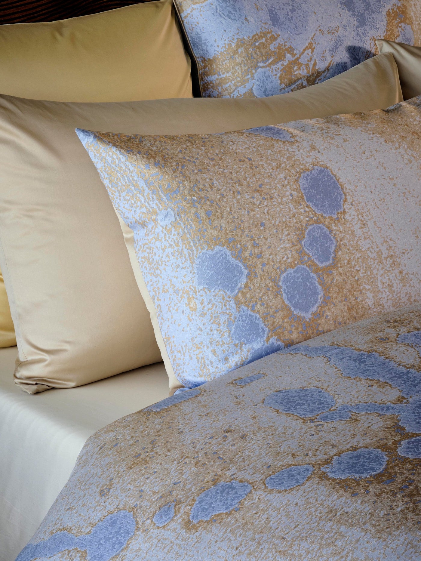 A close-up view of a bed with a light tan silk pillow and a decorative pillow featuring a blue and gold abstract pattern. featuring Callisto Pillowcase by Celso de Lemos available at Linen Society.