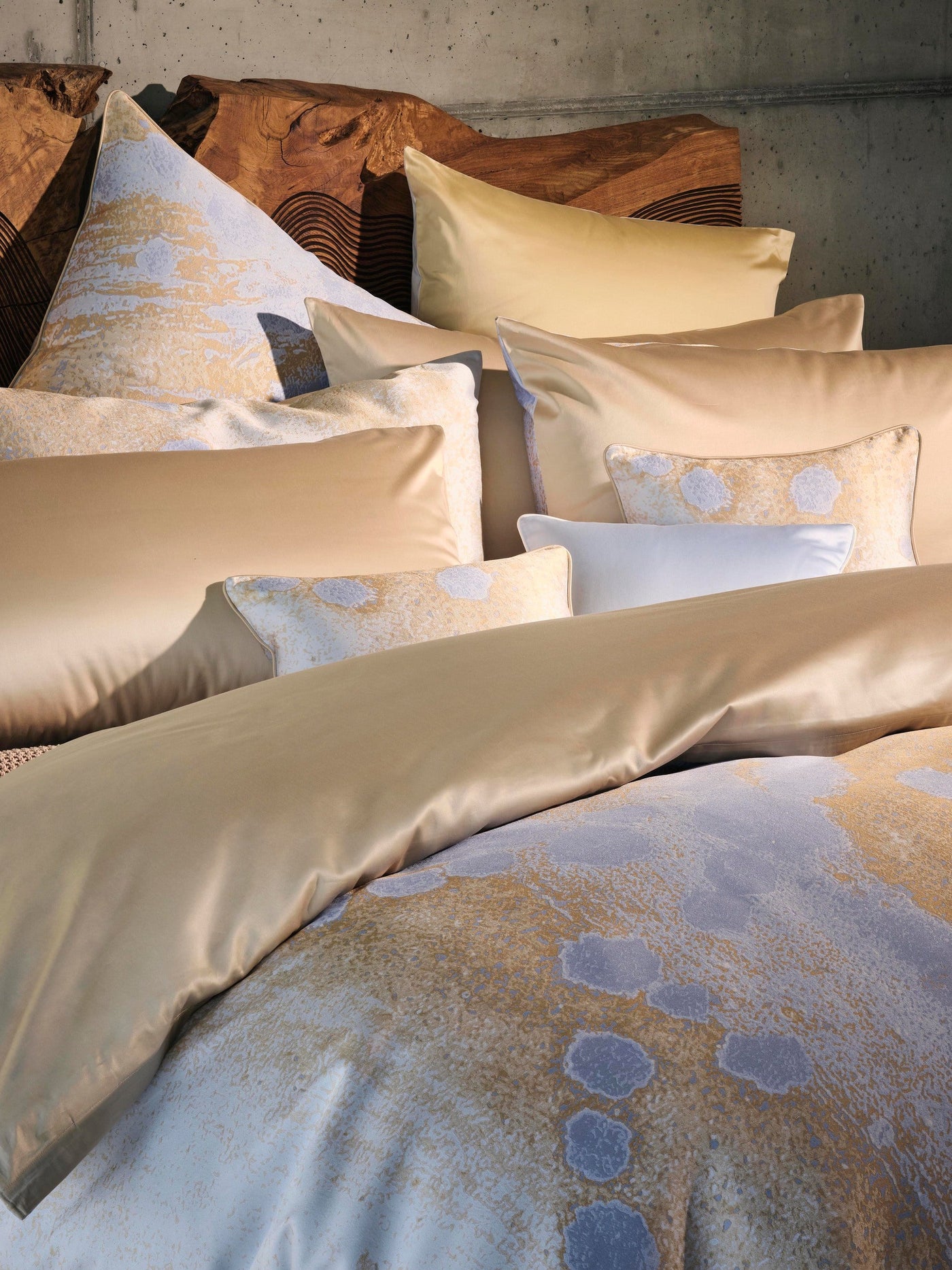 A pile of luxurious, smooth satin pillowcases in golden and pale blue hues, some featuring an abstract gold and grey pattern. featuring Callisto Pillowcase by Celso de Lemos available at Linen Society.