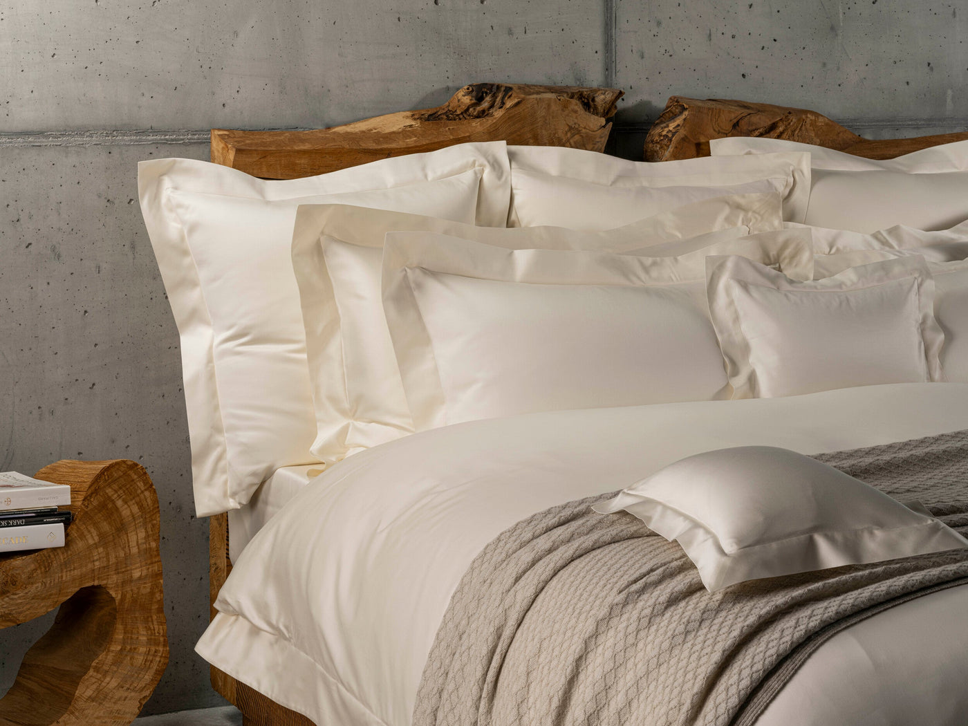 A bed with crisp, off-white pillowcases and a duvet, layered with a textured grey throw, set against a raw concrete wall and rustic wooden headboard, featuring Capela Pillowcase by Celso de Lemos available at Linen Society.