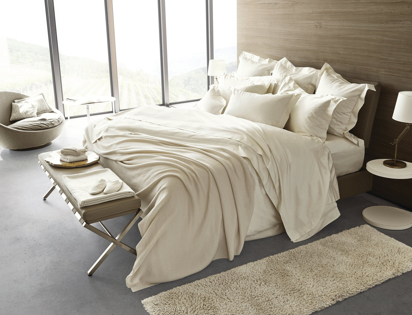 A bed draped with light cream bedding and a textured throw, styled in a modern bedroom with large windows. featuring Capela Pillowcase by Celso de Lemos available at Linen Society.