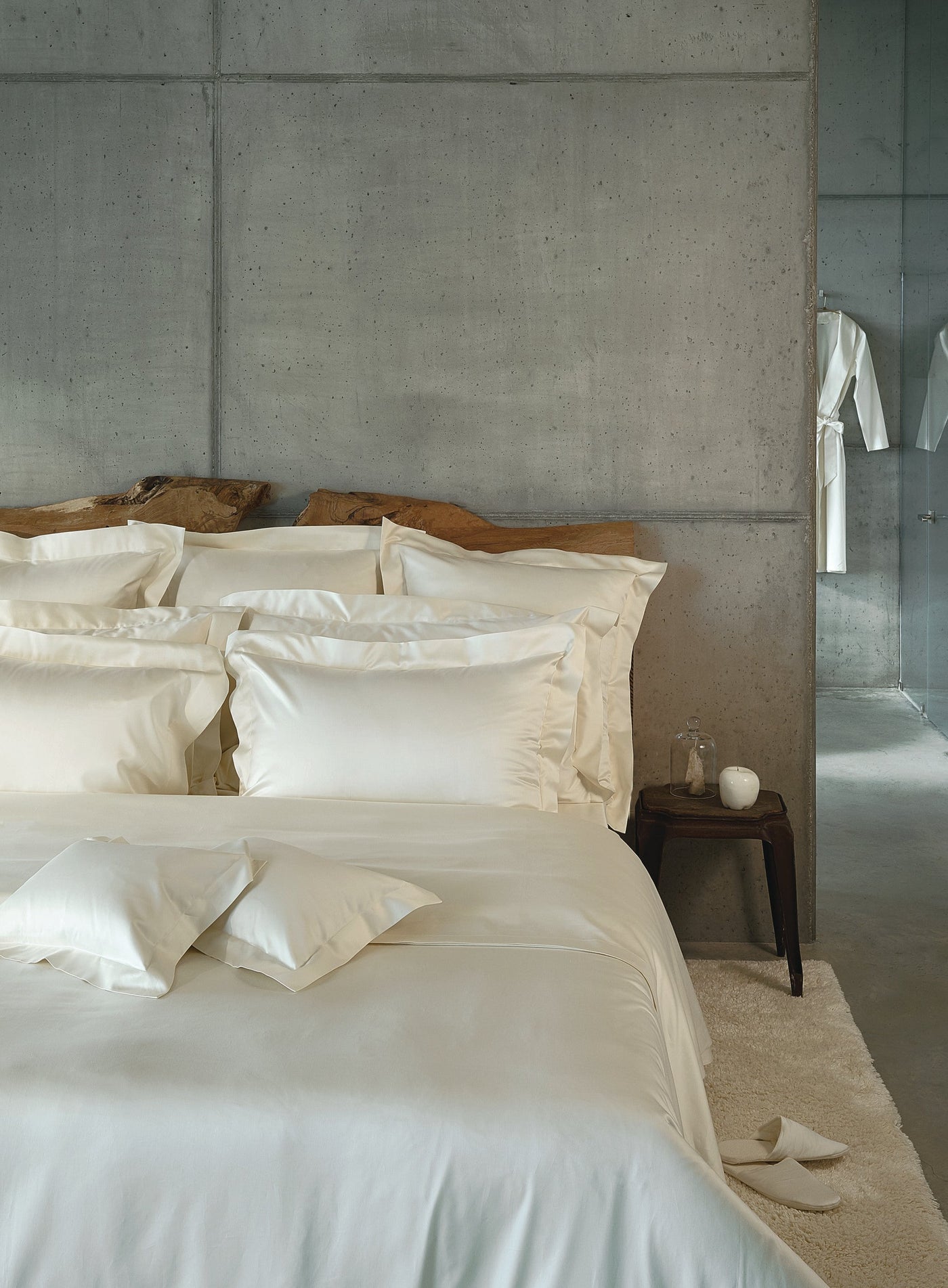 A bed with white bedding is set against a concrete wall and a rustic wooden headboard. featuring Capela Pillowcase by Celso de Lemos available at Linen Society.