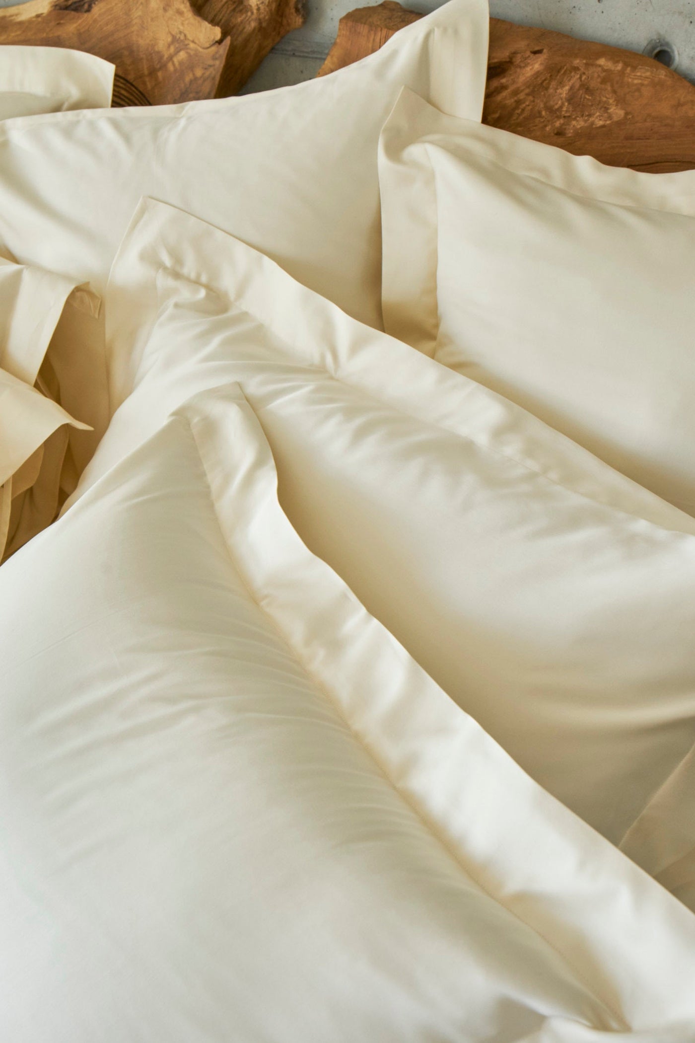 Soft, cream-colored bedding is layered and rumpled, showcasing a smooth texture. featuring Capela Pillowcase by Celso de Lemos available at Linen Society.