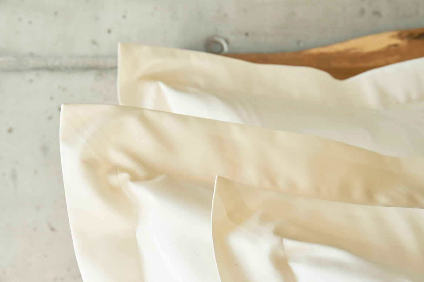Cream-colored pillowcases are layered with soft folds against a concrete background, featuring the Capela Pillowcase by Celso de Lemos available at Linen Society.