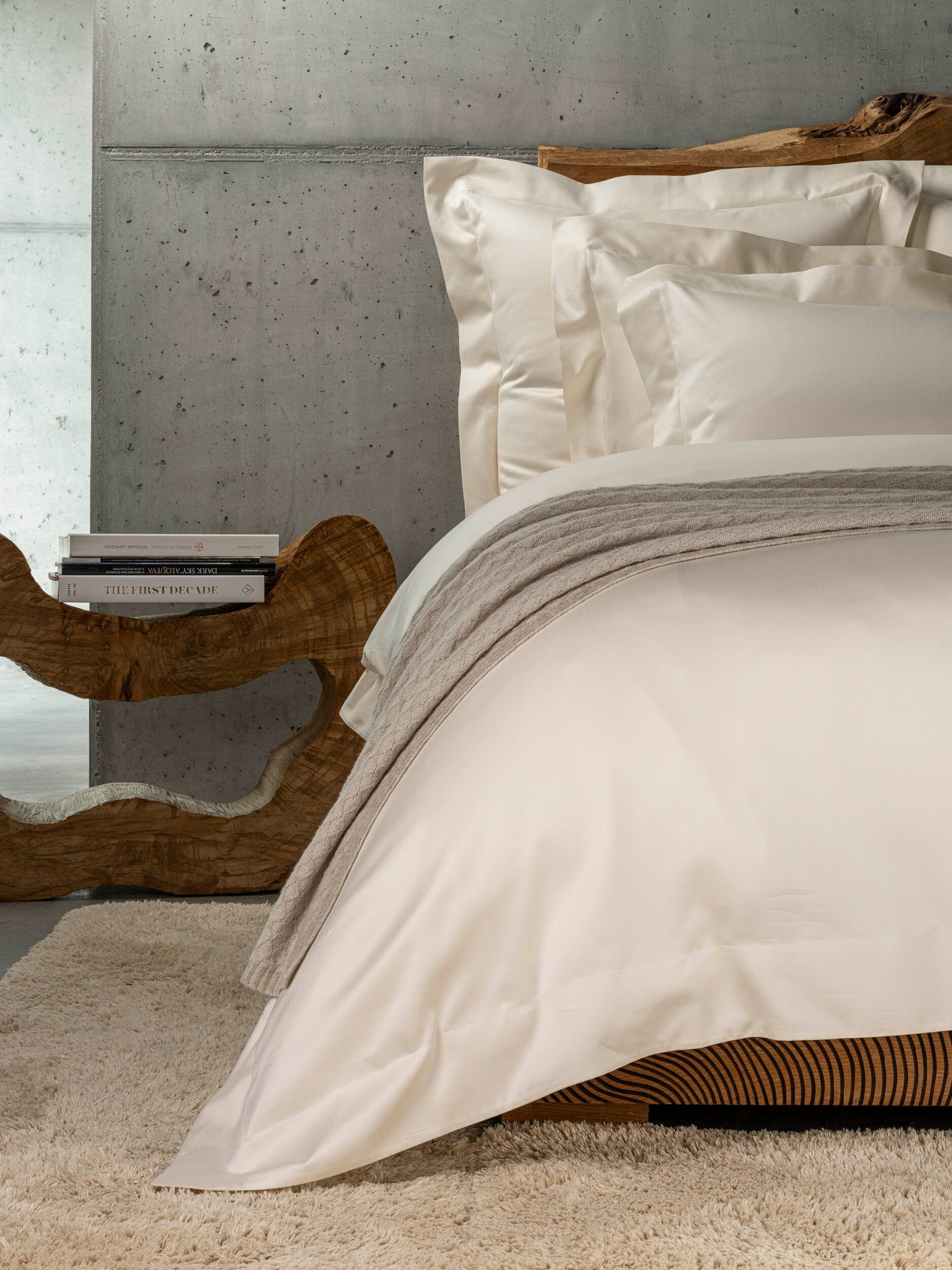 Close-up of crisp white pillowcases folded neatly on a bed with a grey knit throw. featuring Capela Pillowcase by Celso de Lemos available at Linen Society.