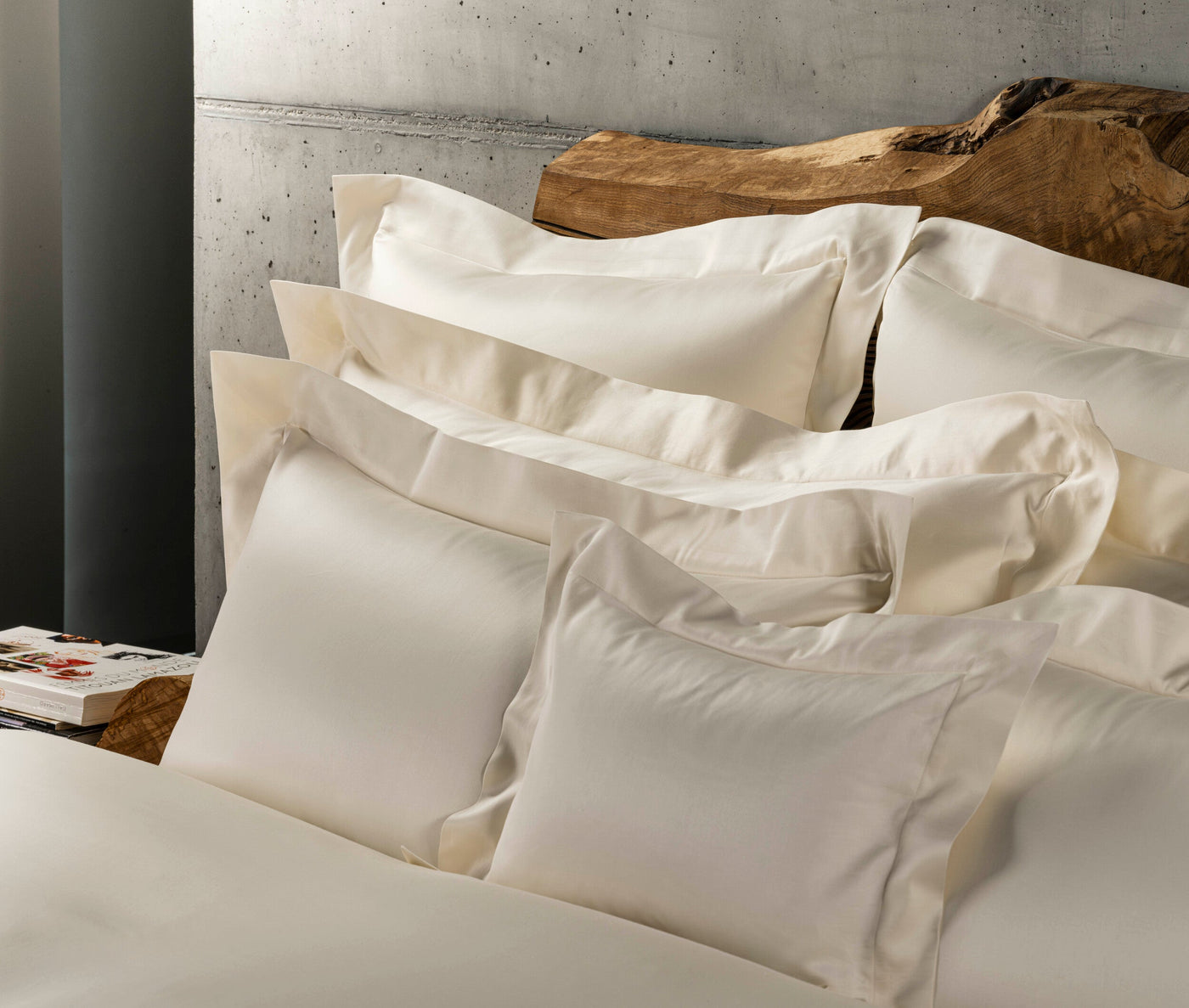 Close-up of soft, off-white bedding arranged against a textured concrete wall and a live-edge wooden headboard, featuring Capela Pillowcase by Celso de Lemos available at Linen Society.
